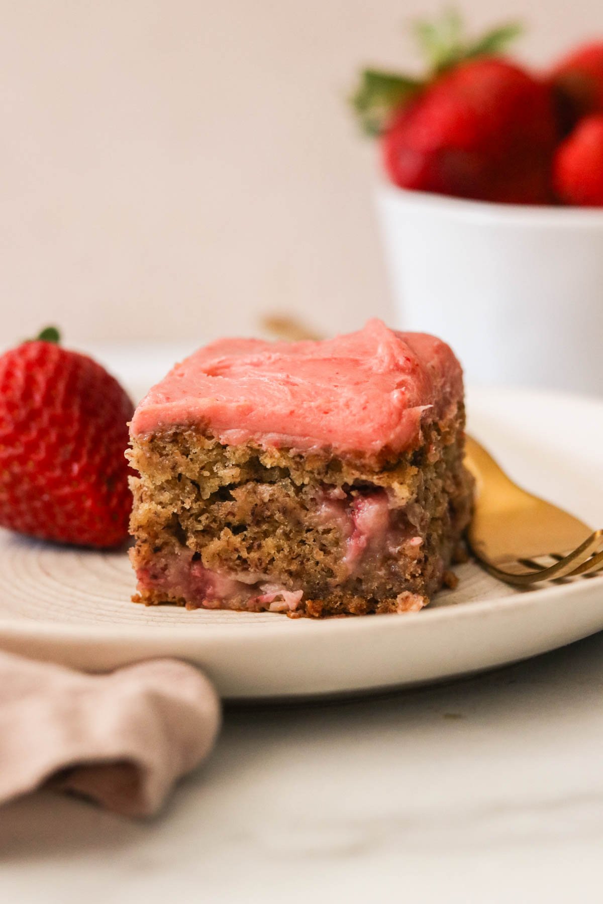a slice of strawberry banana cake on a white plate with a gold fork next to a strawberry.