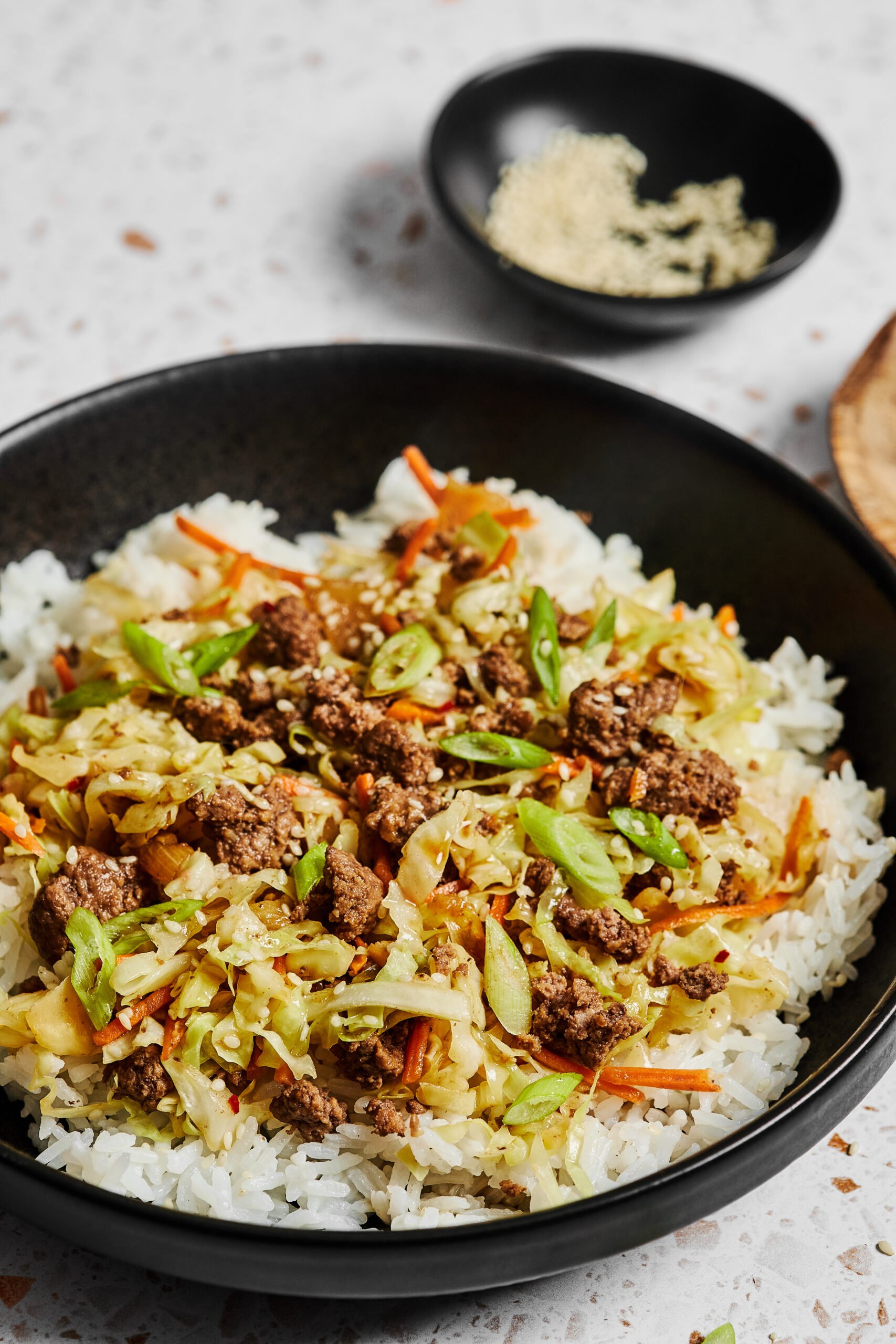 Close-up of Asian ground beef and cabbage stir fry served over white rice in a black bowl on a white speckled countertop.