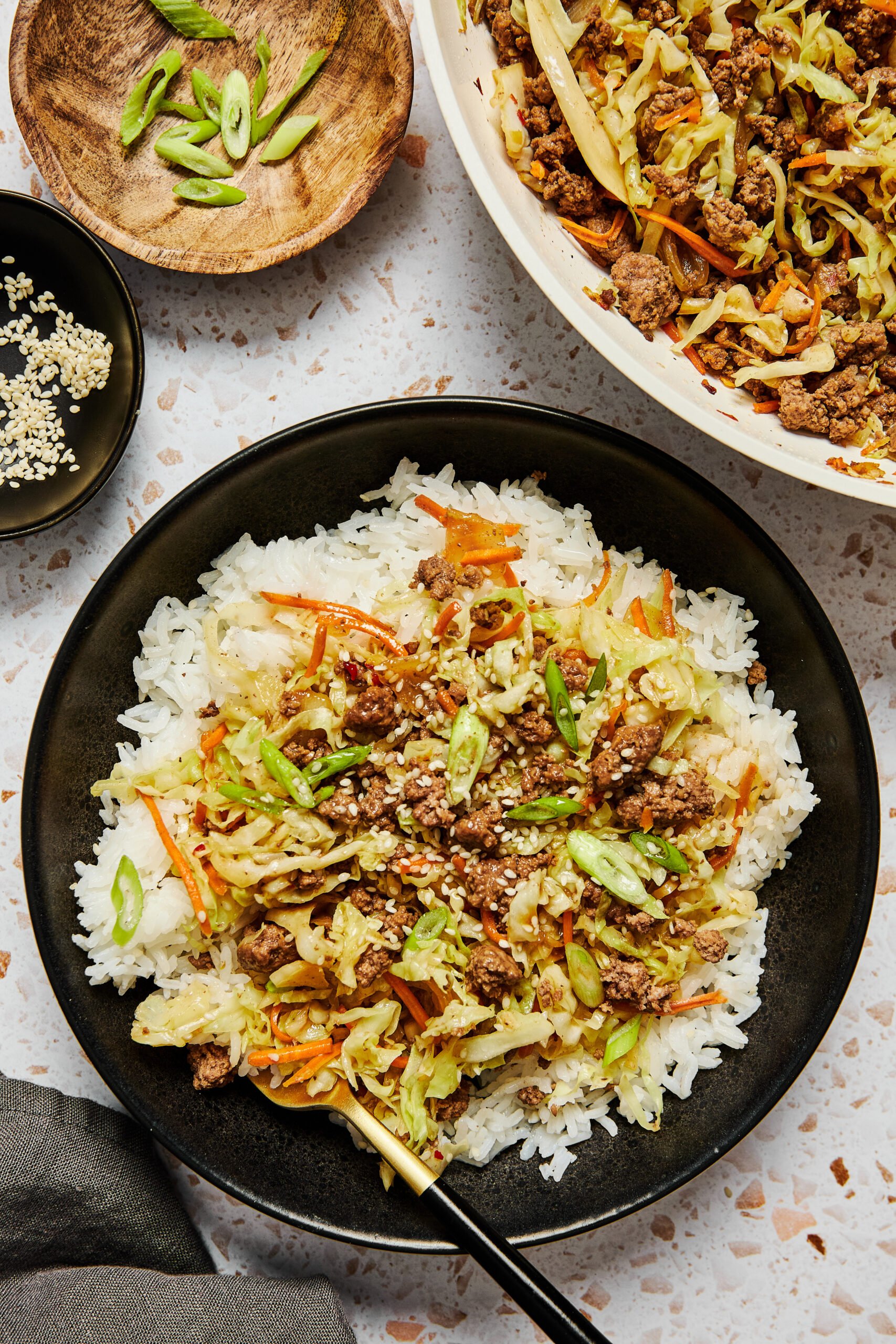 Asian ground beef and cabbage stir fry over rice in a black bowl, with a pan of stir fry, sesame seeds in a small bowl, and chopped green onions in a wooden bowl.