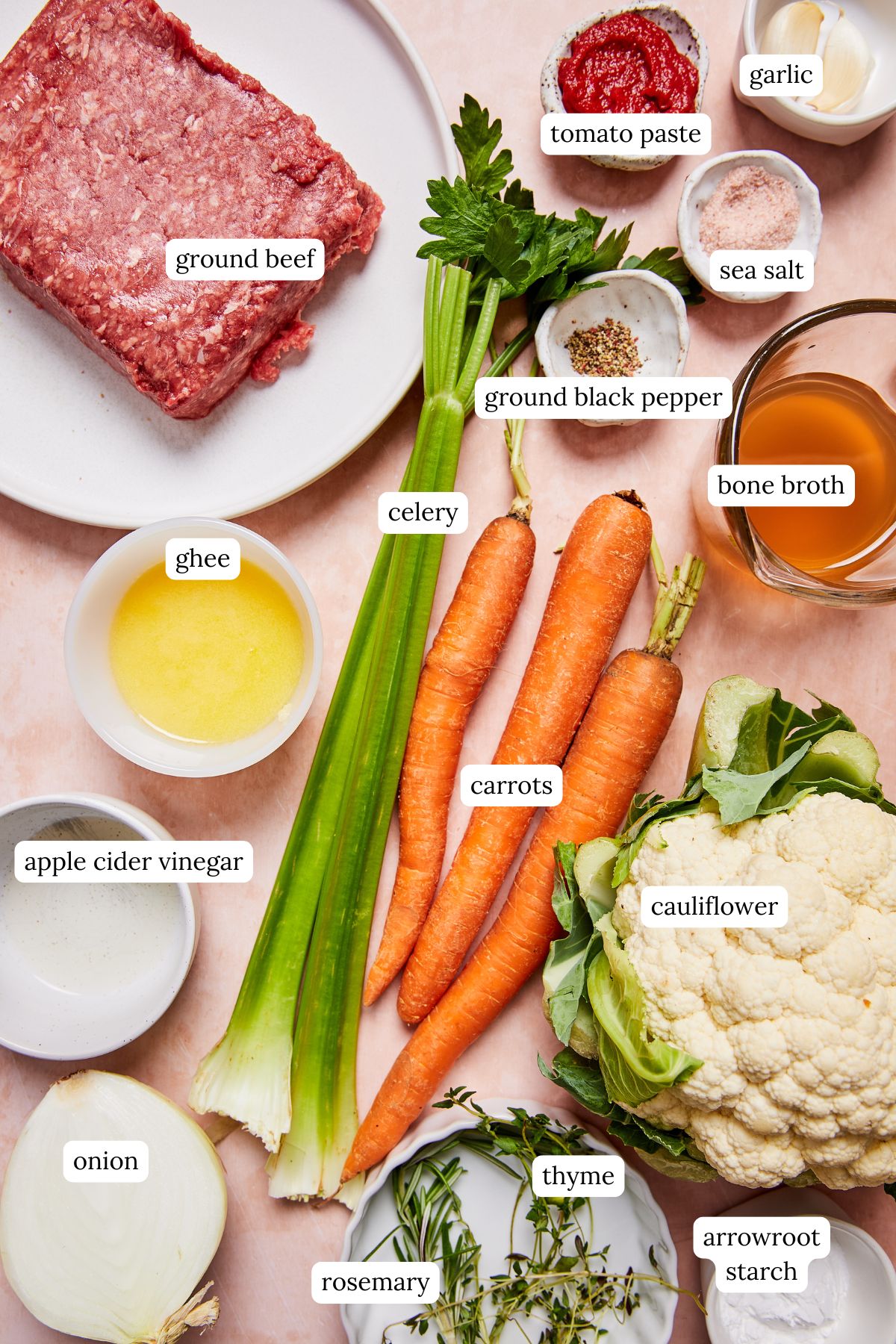 Ingredients for low-carb shepherd’s pie arranged in bowls, including cauliflower, carrots, ground meat, onion, garlic, celery, herbs, broth, and vinegar on a light pink surface.