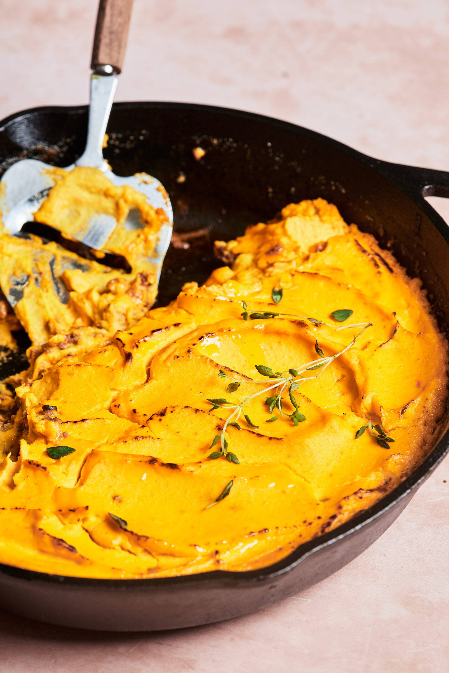 Cast iron skillet with low-carb shepherd’s pie, baked with a golden cauliflower and carrot mash topping, half eaten with a spatula resting inside.