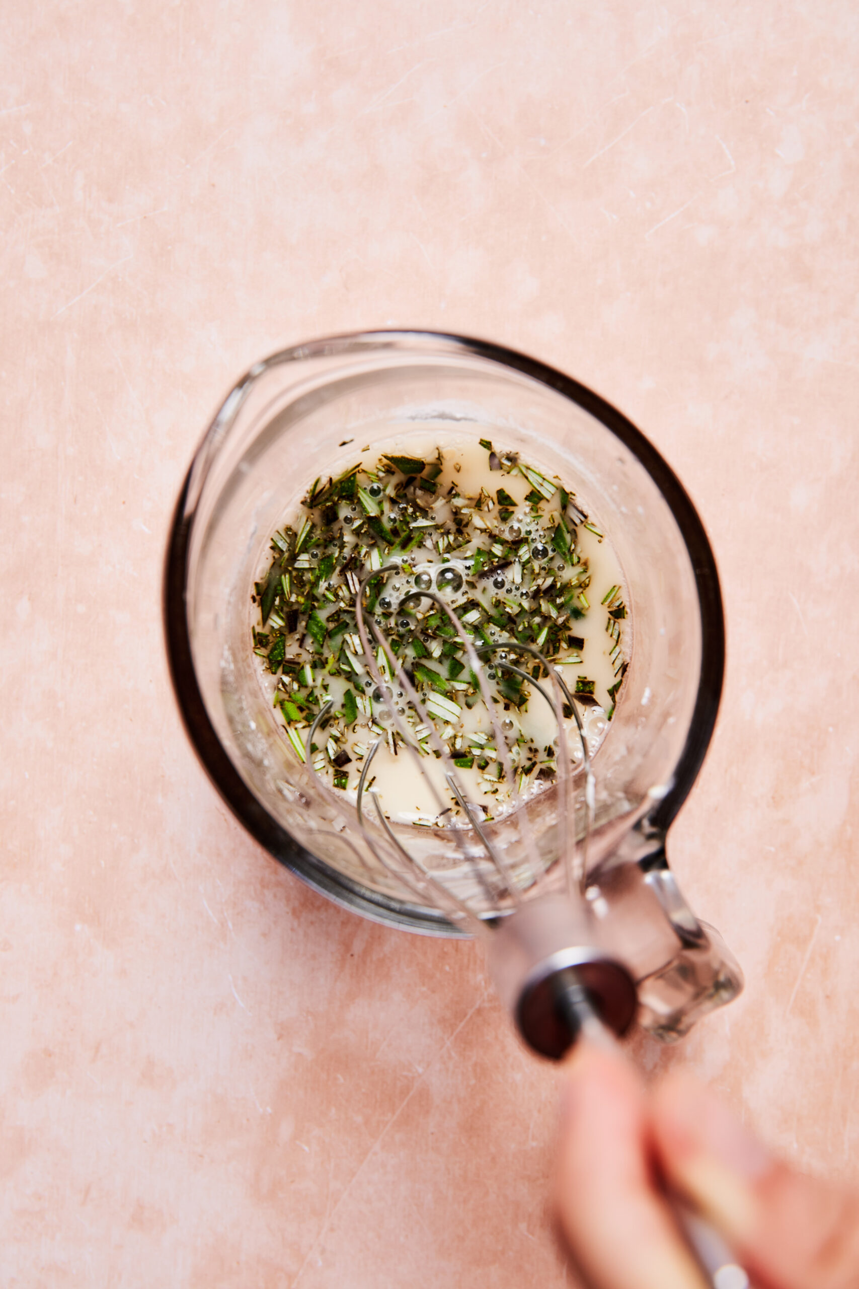 A hand whisking the  broth mixture with vinegar and herbs in a glass measuring cup on a light pink surface.