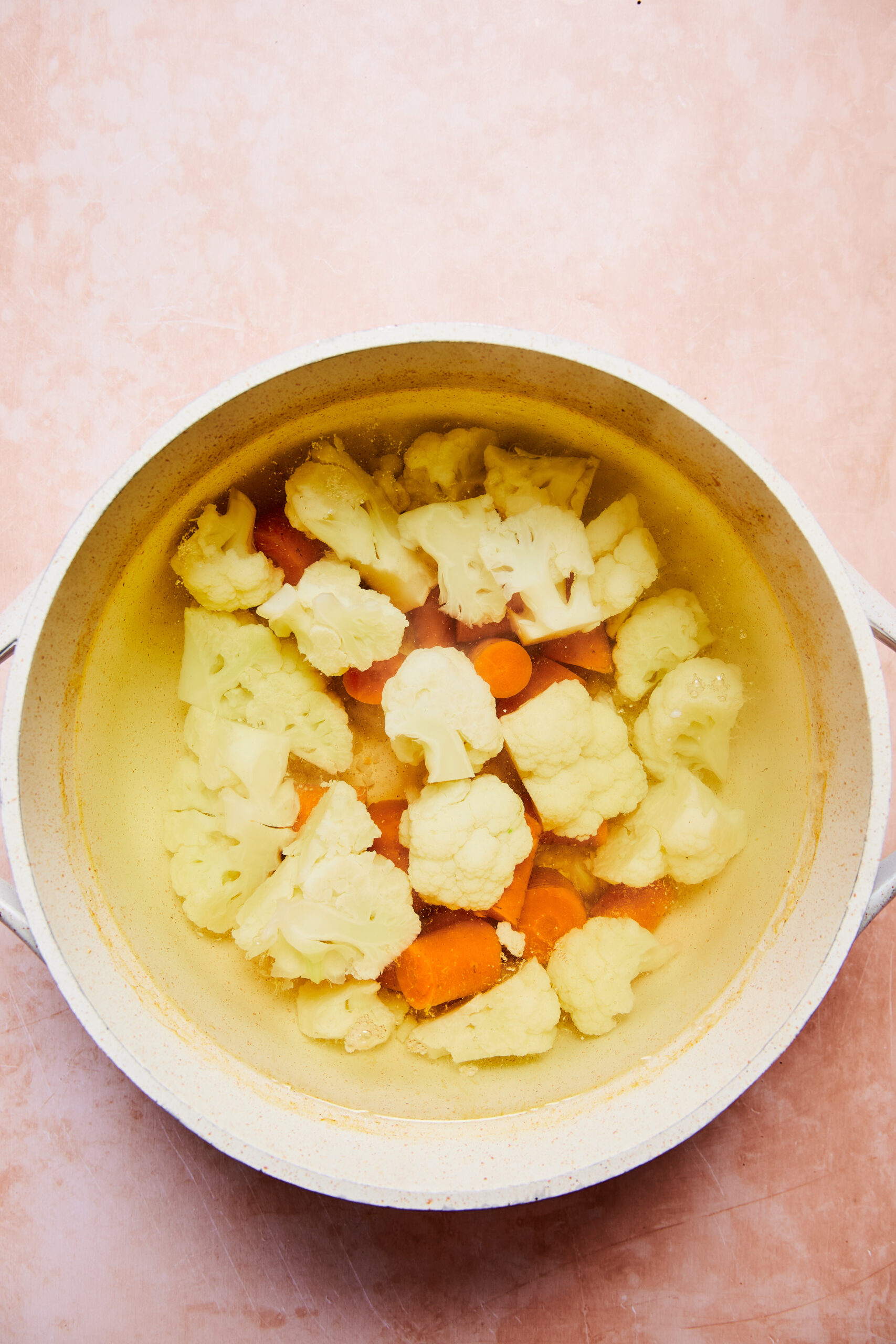 Carrots and cauliflower in a pot of water on a light pink surface.