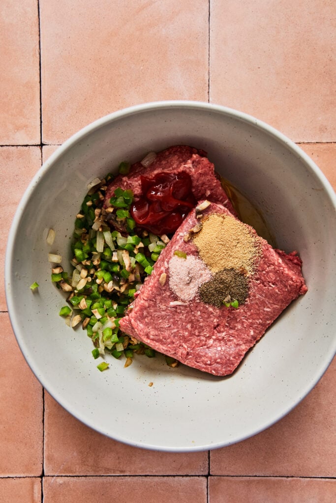 Raw ground beef, sautéed vegetables, eggs, and seasonings combined in a glass mixing bowl for meatloaf mixture.
