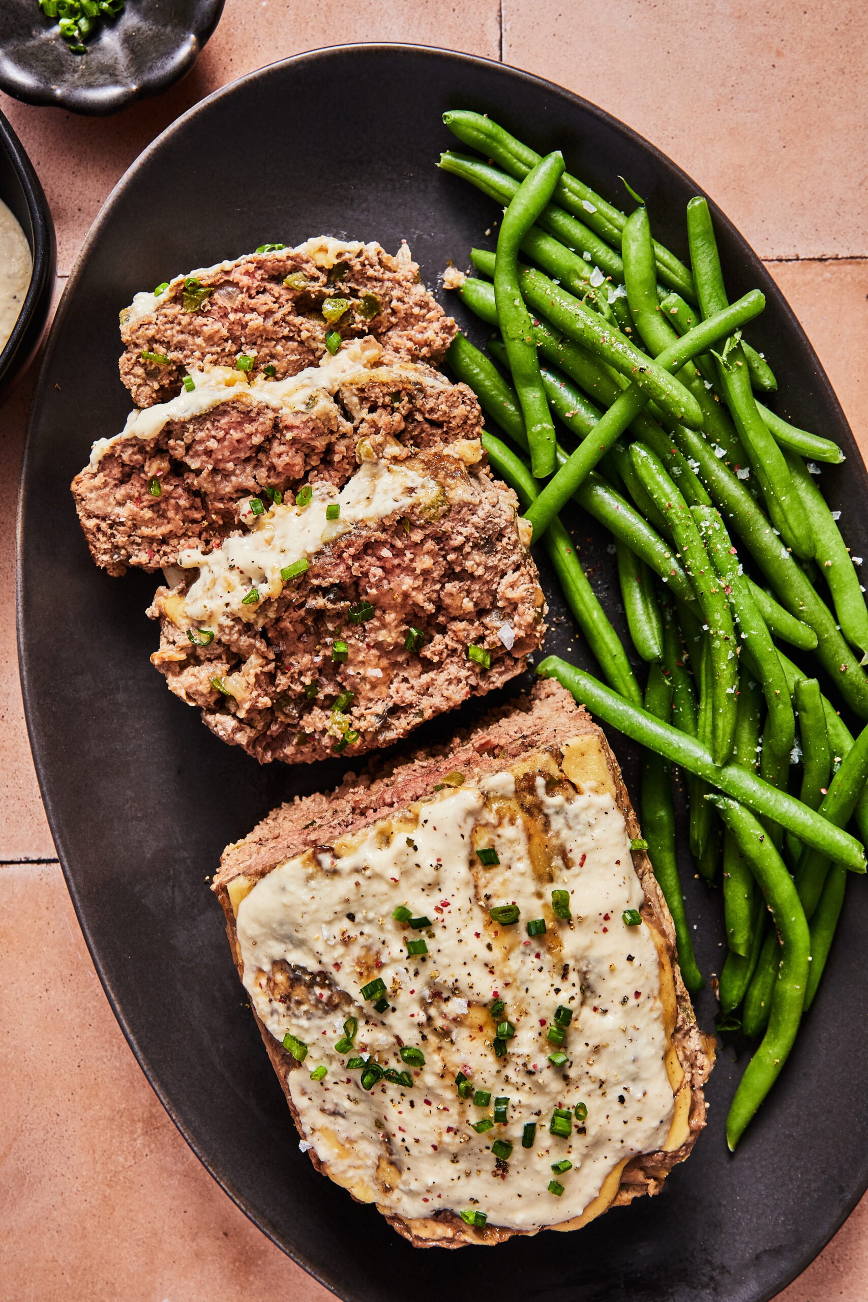 Sliced Philly cheesesteak meatloaf topped with creamy cashew cheese sauce on a black platter with a side of seasoned green beans.