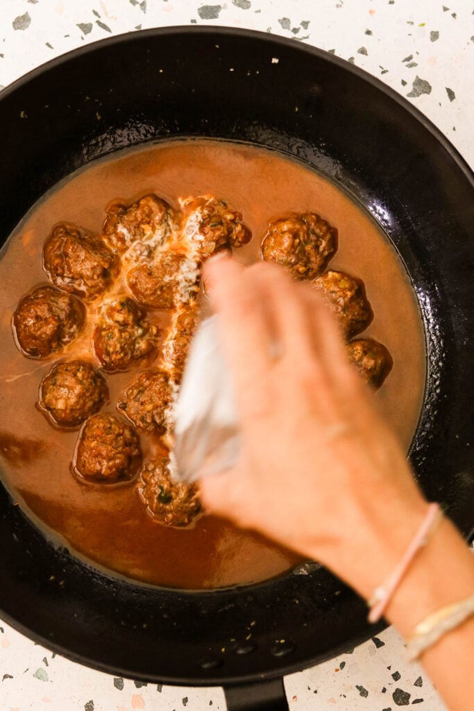 a hand pouring the slurry into the pan of meatballs and sauce. 