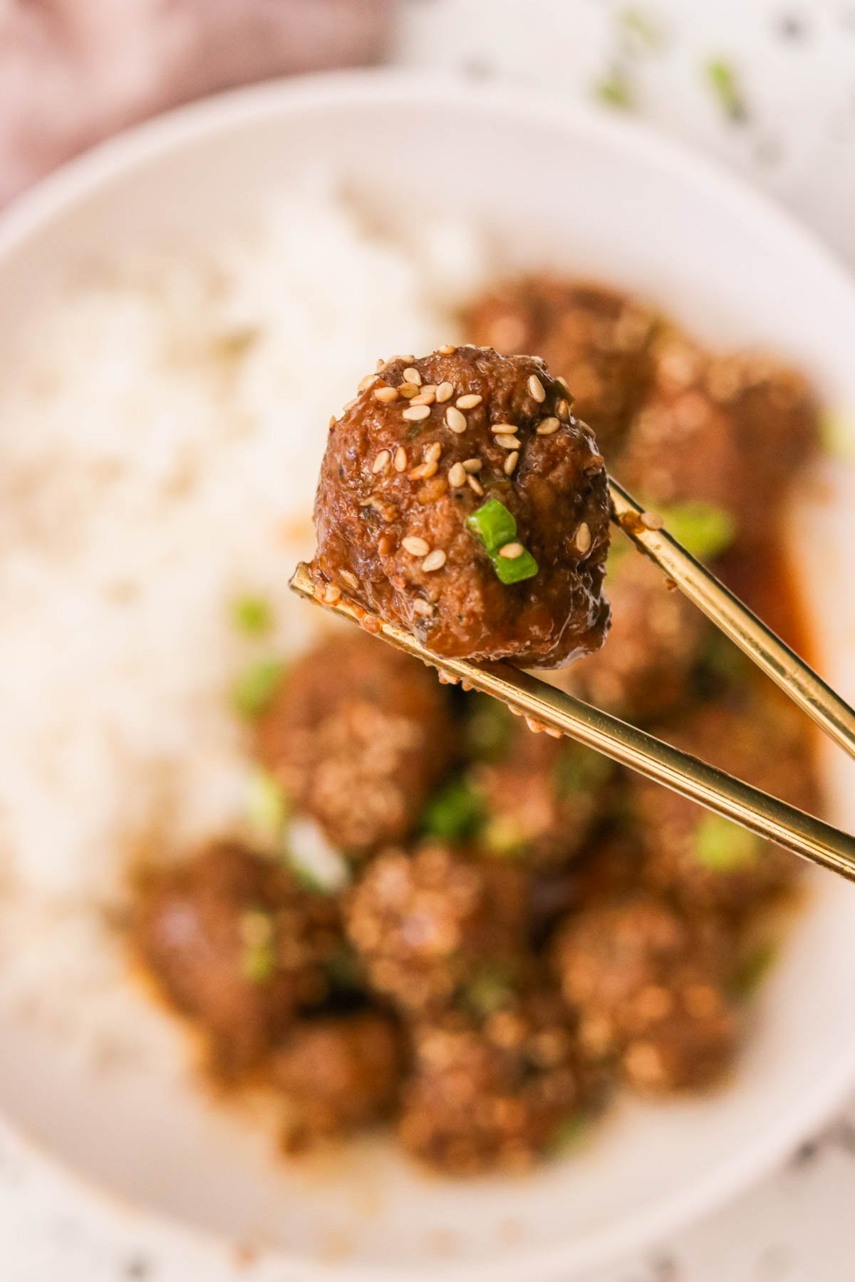 gold chopsticks holding a single meatball covered in sauce and garnished with sesame seeds and green onions