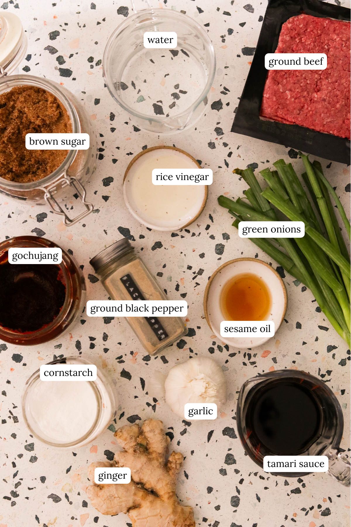 recipe ingredients on a speckled surface including brown sugar, rice vinegar, ground beef, sesame oil, gochujang, cornstarch, black pepper, garlic, ginger, tamari, and water