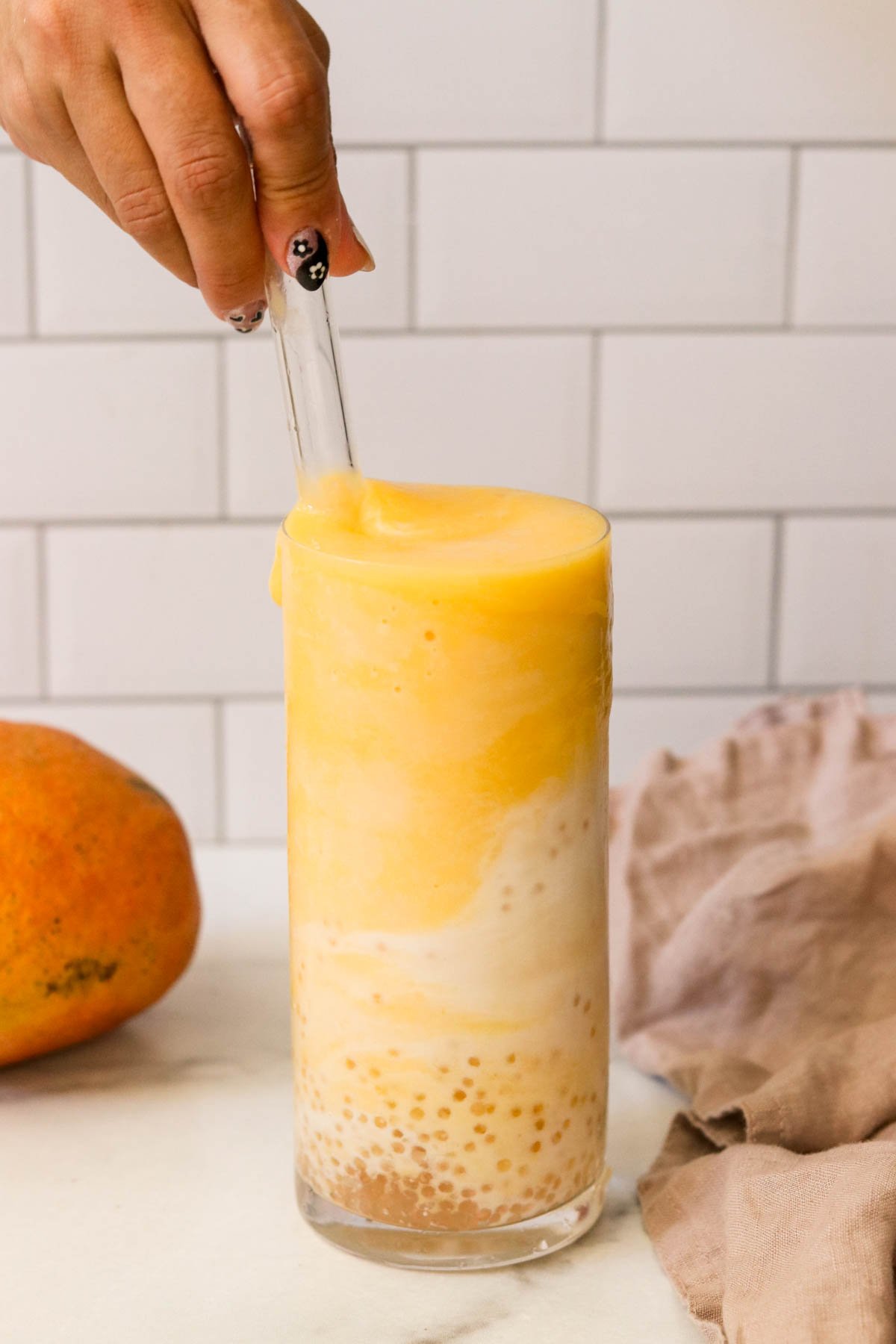 A hand stirring a glass straw in a tall glass of mango sticky rice smoothie in front of a light colored background.