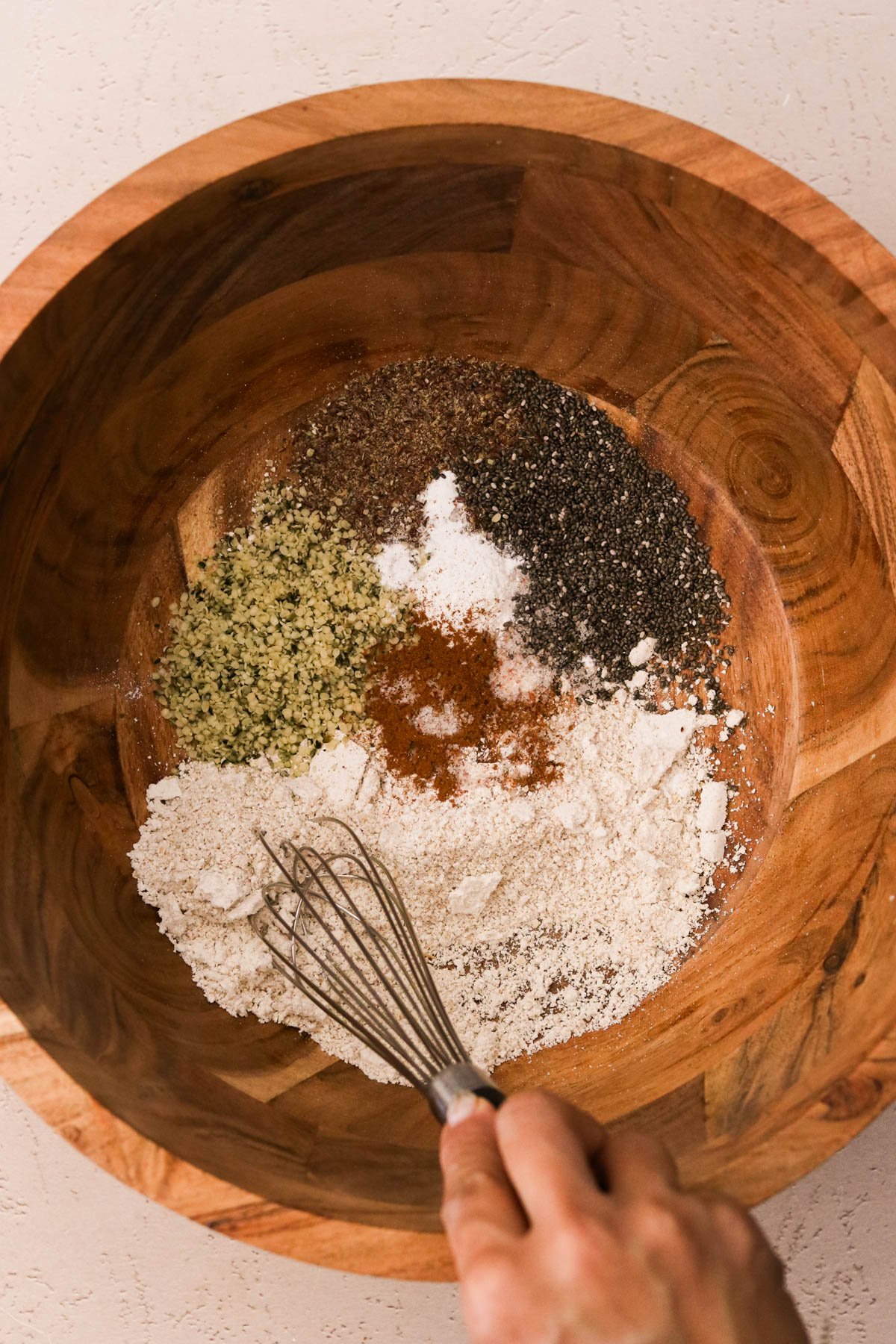 A hand whisking oat flour, flaxseed, chia seeds, hemp, cinnamon, baking powder, and salt together in a wooden mixing bowl.