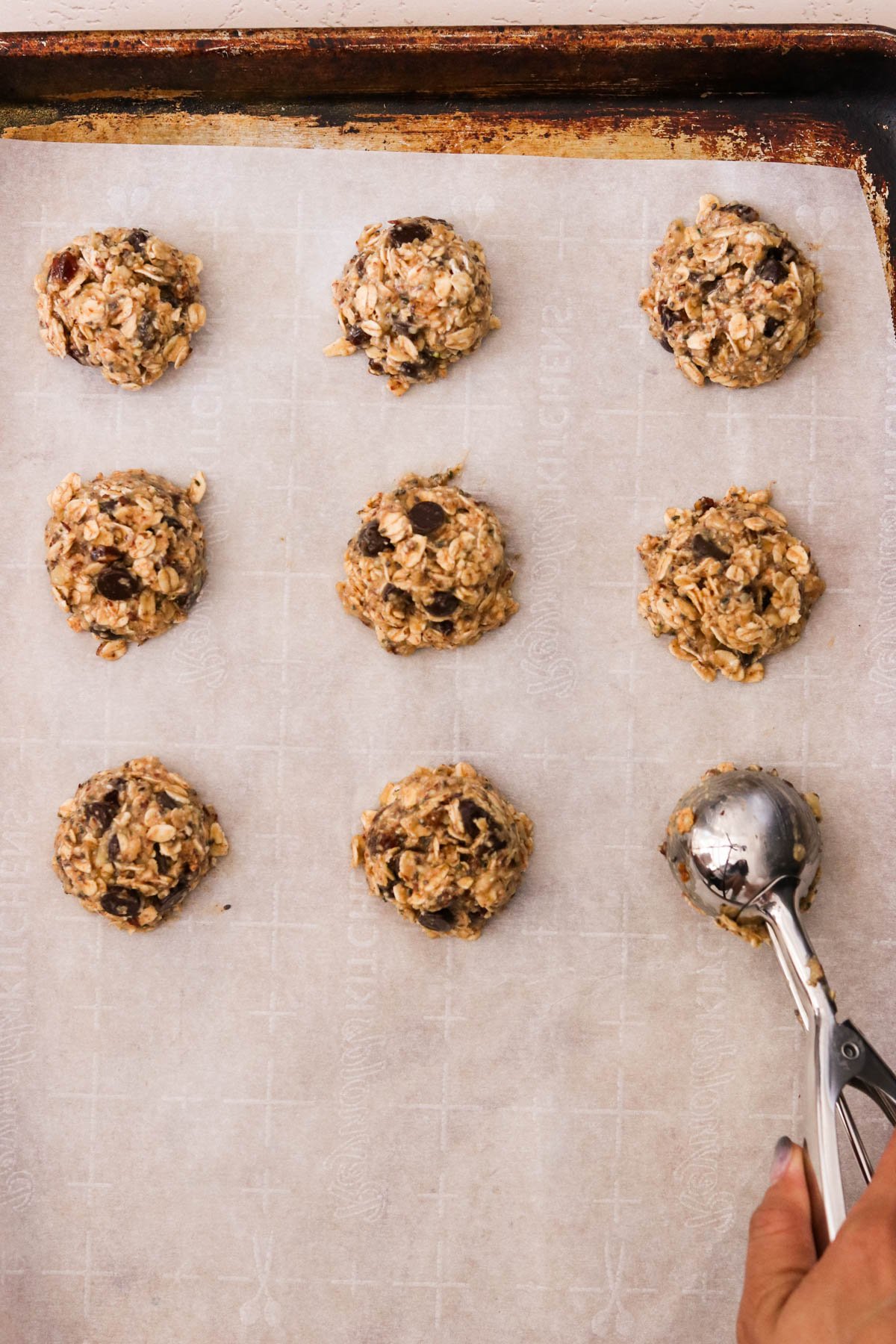 A silver cookie scoop dropping breakfast oatmeal cookie dough onto a parchment-lined baking sheet.
