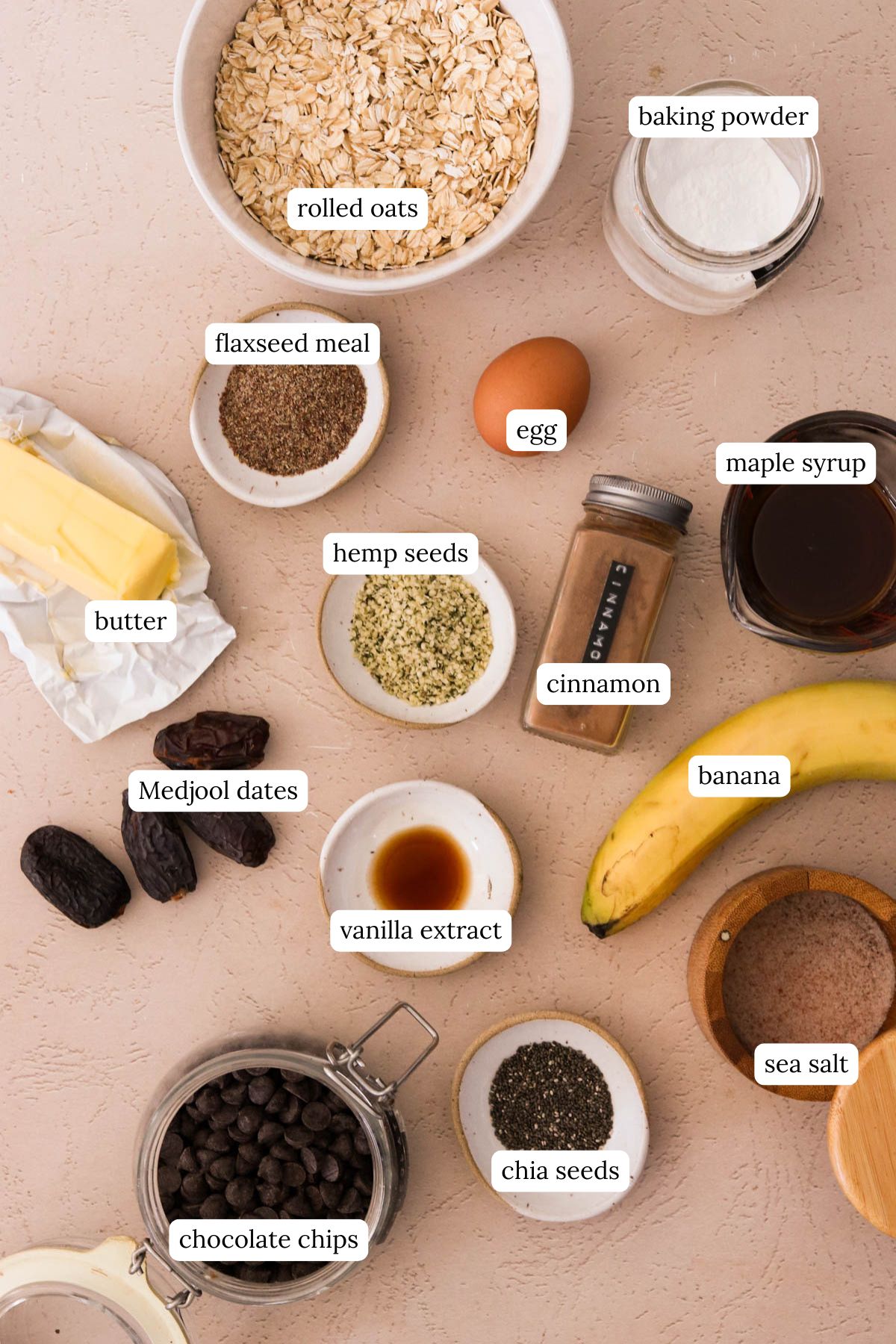 Ingredients for oatmeal breakfast cookies including rolled oats, flaxseed meal, chia, hemp seeds, banana, and maple syrup on a light pink background.