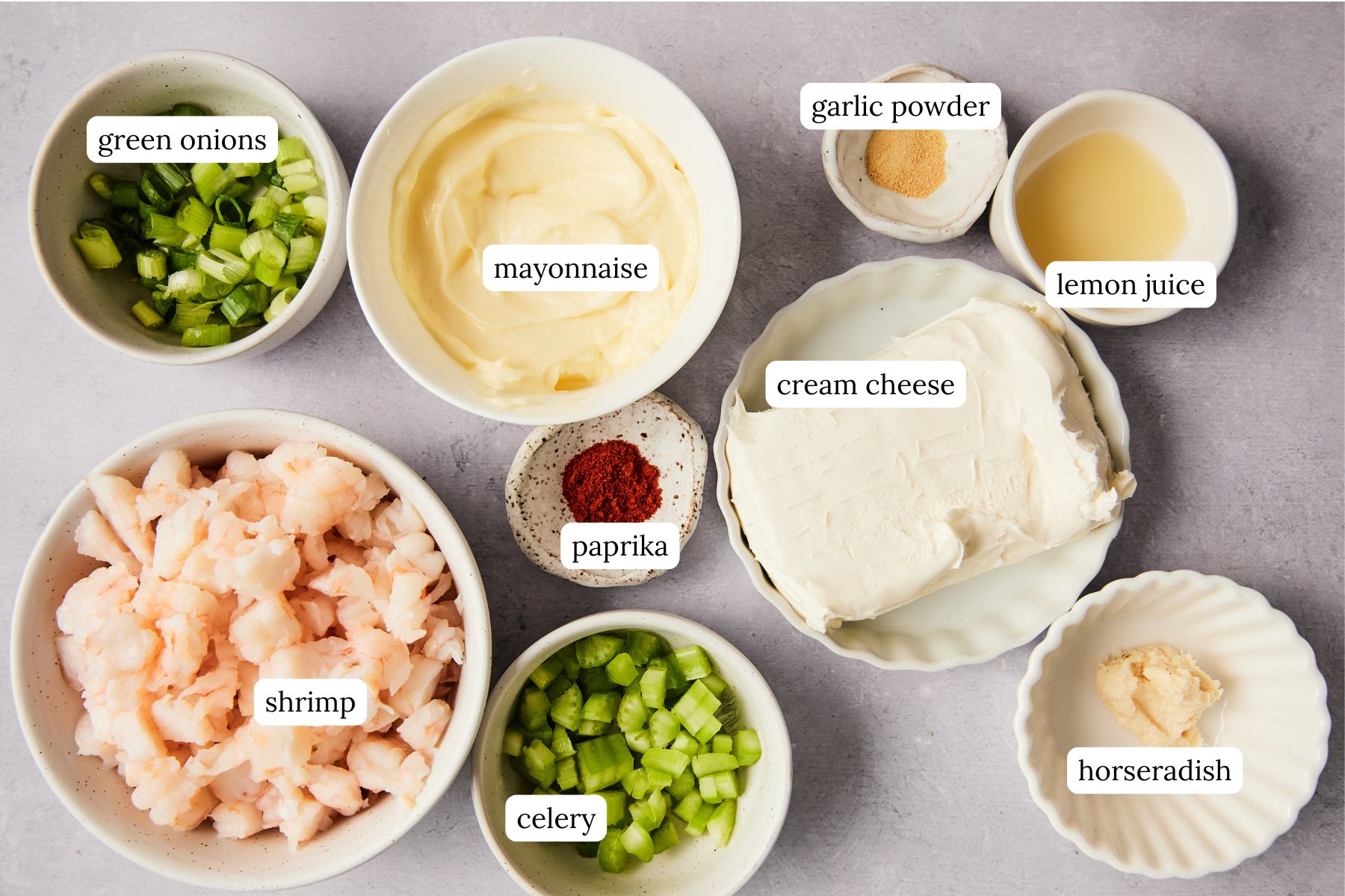 Ingredients for shrimp dip arranged on a counter including cream cheese, mayonnaise, lemon, horseradish, paprika, garlic powder, chopped shrimp, celery, and green onions.