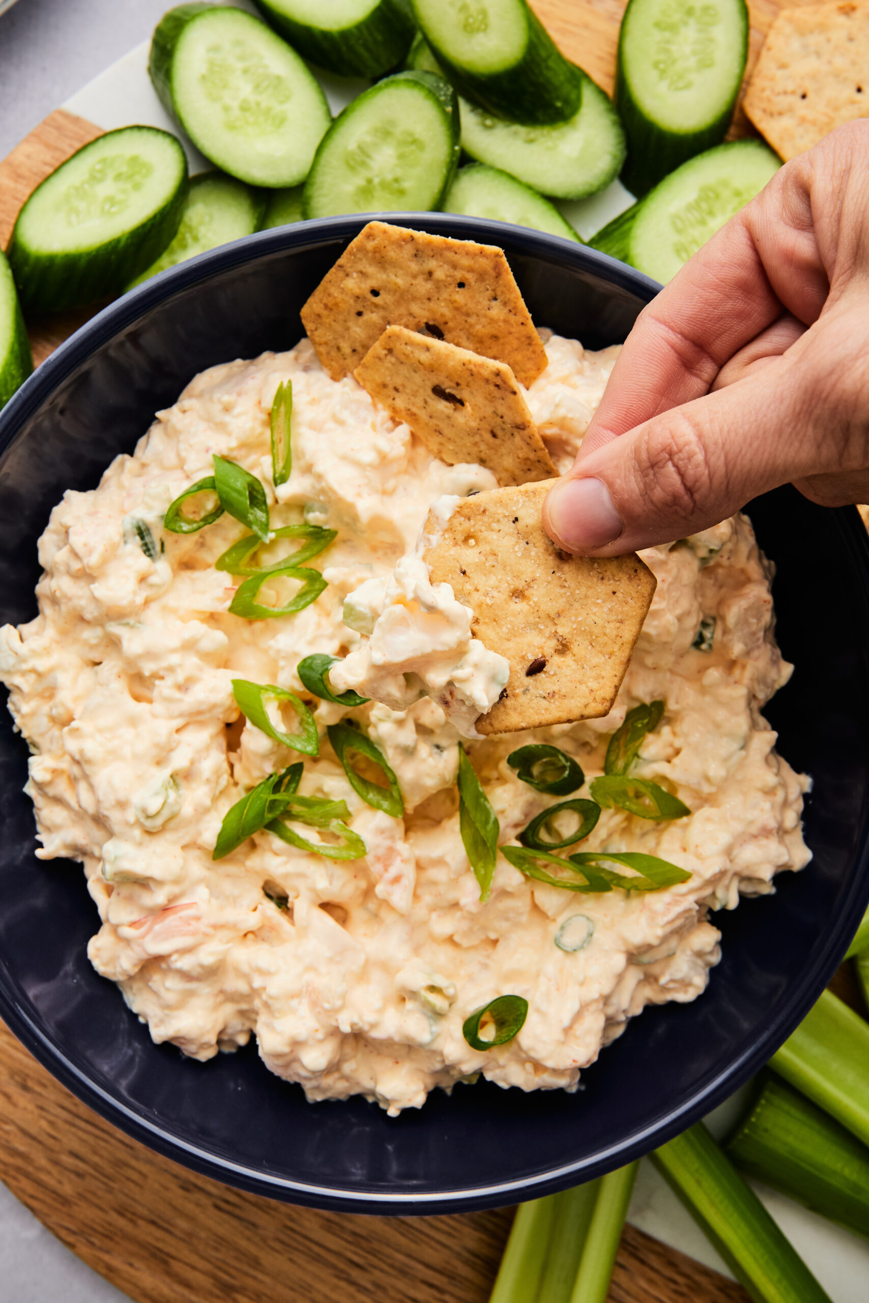 Close-up of a hand dipping a cracker into creamy shrimp dip garnished with green onion.