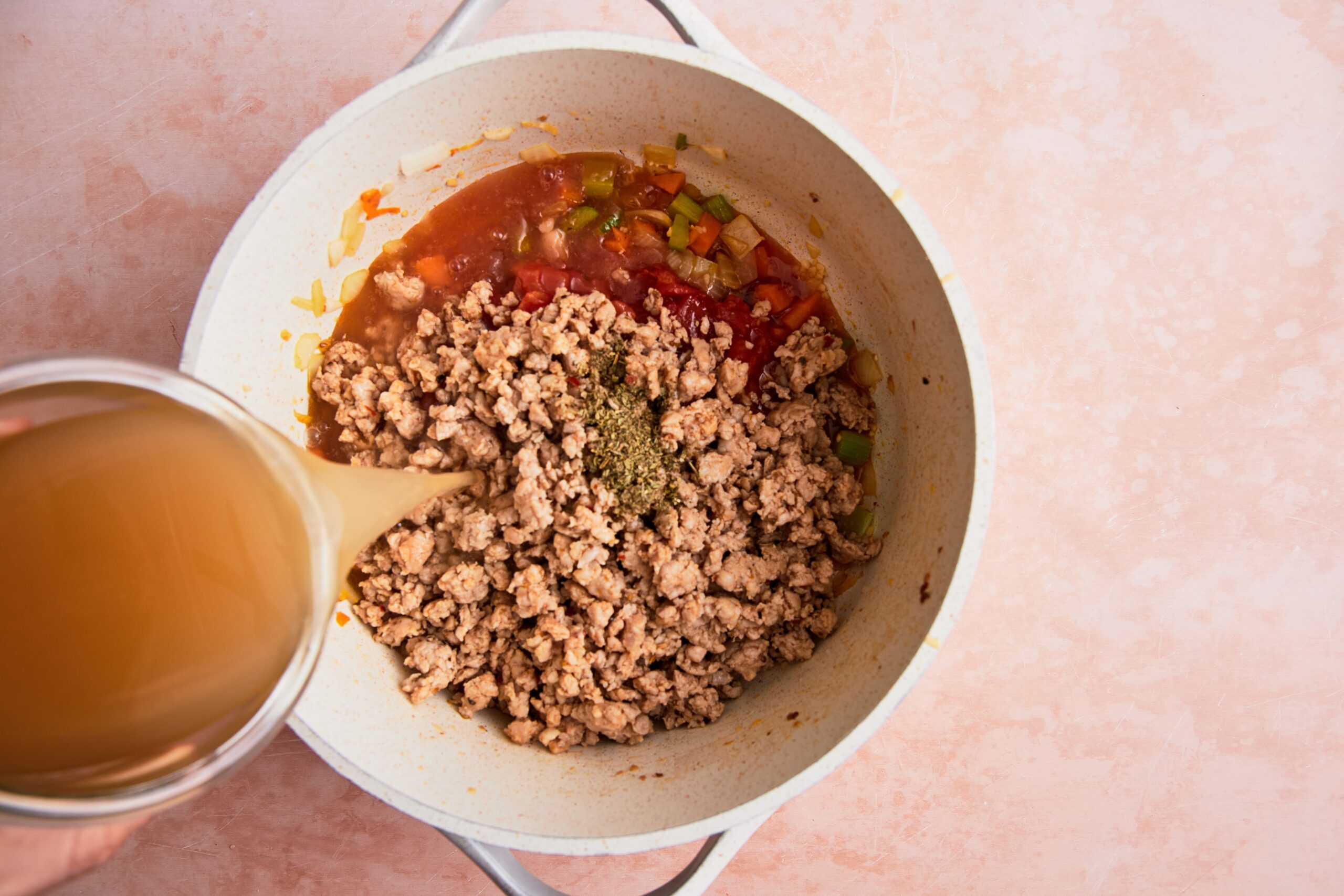 Broth being poured into a large white soup pot with tomatoes, seasoning, and cooked sausage.