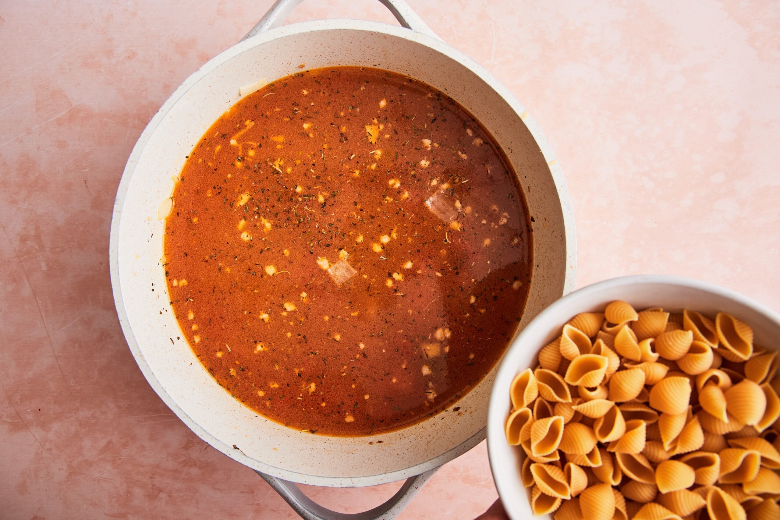 Uncooked shell pasta being added to boiling tomato broth with sausage in a soup pot.
