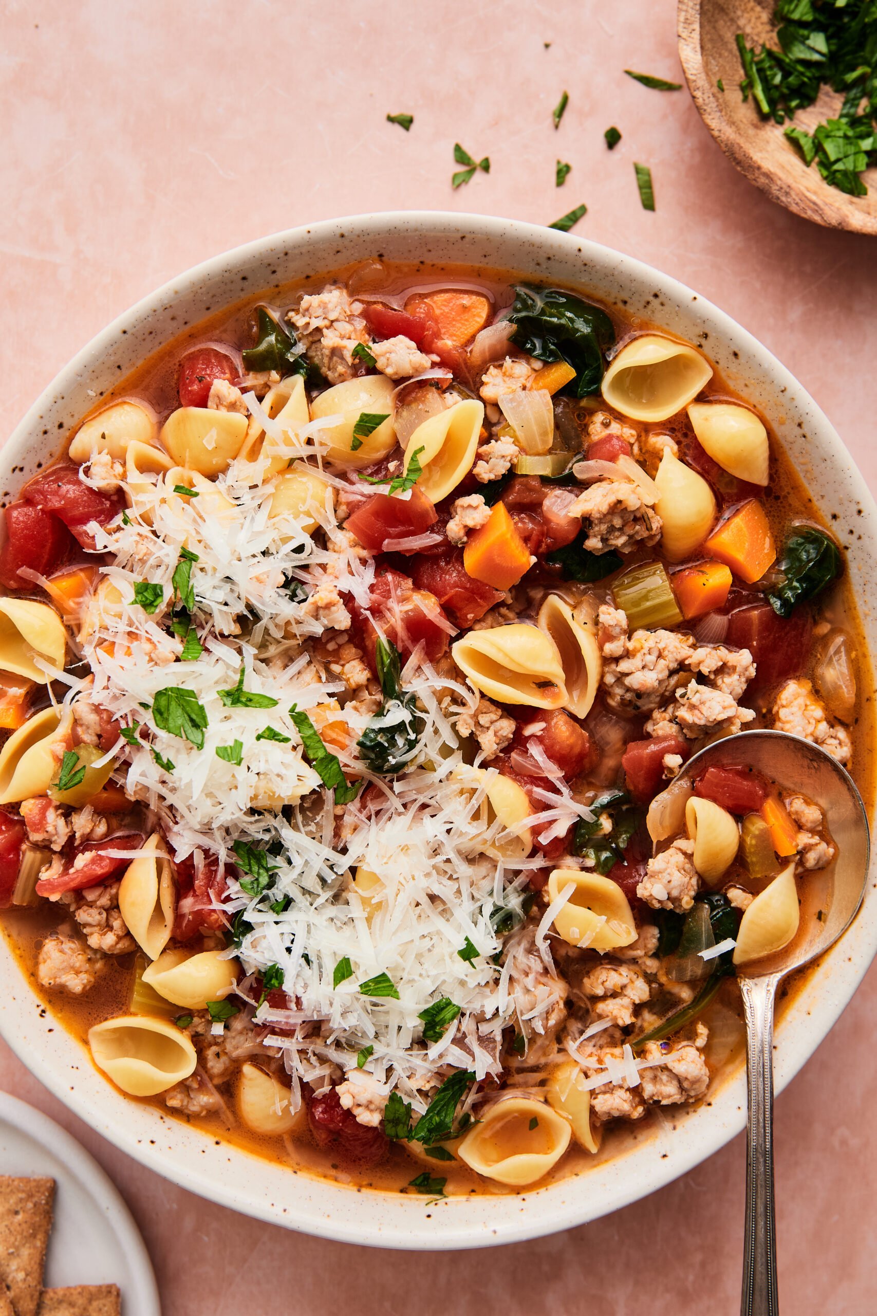 A bowl of hearty Italian sausage and pasta soup filled with shell pasta, spinach, tomatoes, carrots, and sausage, served with a spoon on the side.