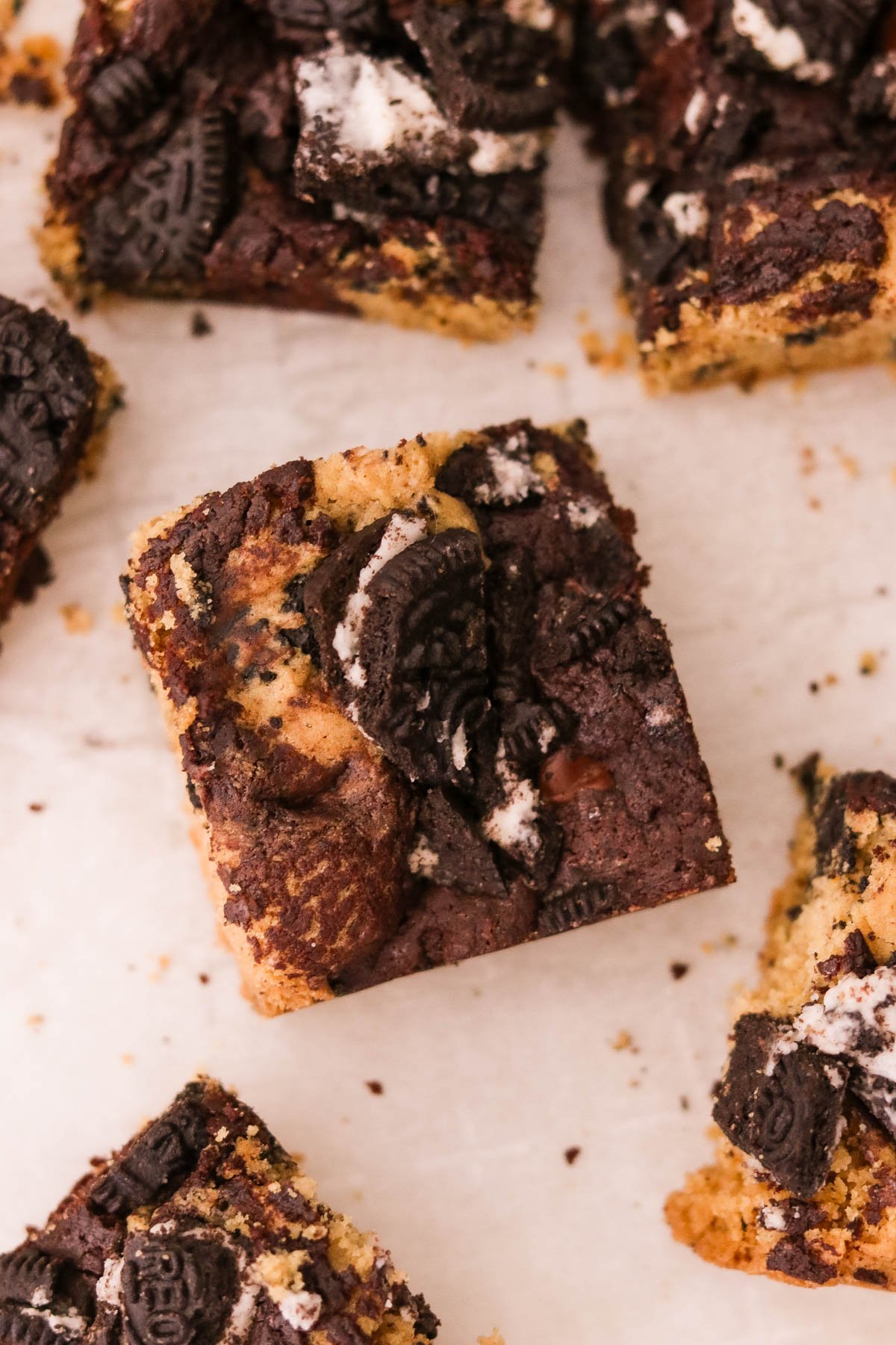 Close-up of a fudgy Oreo brookie square topped with chopped Oreos on parchment paper, with more brookie squares scattered in the background.