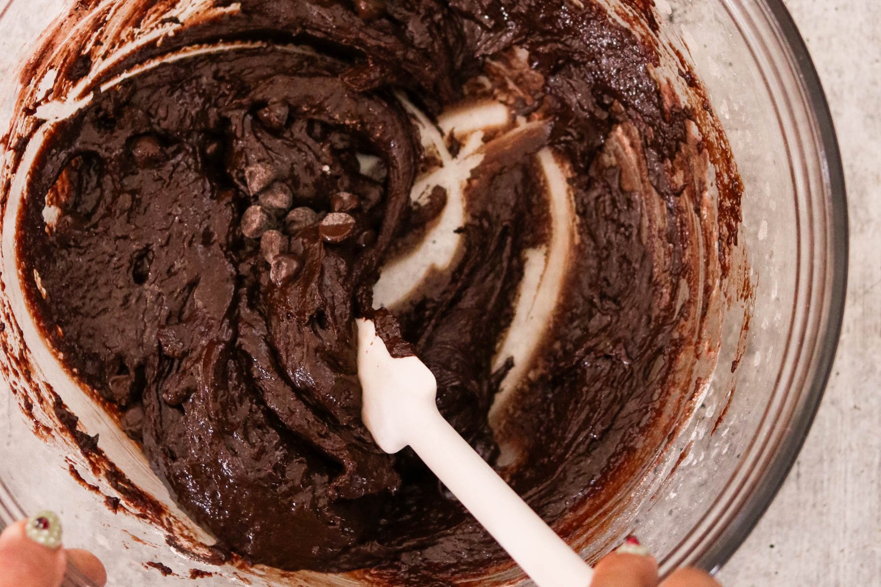 Glass bowl of brownie batter with a spatula mixing the chocolate batter.
