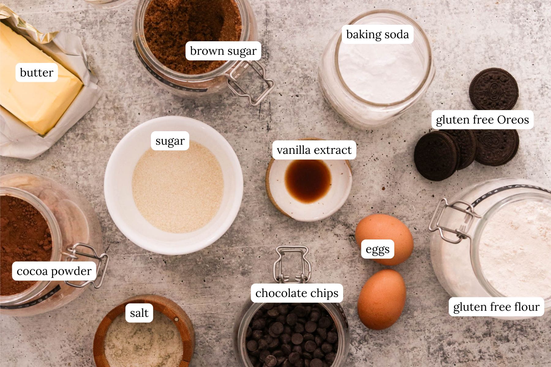 Overhead view of Oreo brookies ingredients including chocolate chips, flour, sugar, butter, eggs, and Oreos on light gray counter.