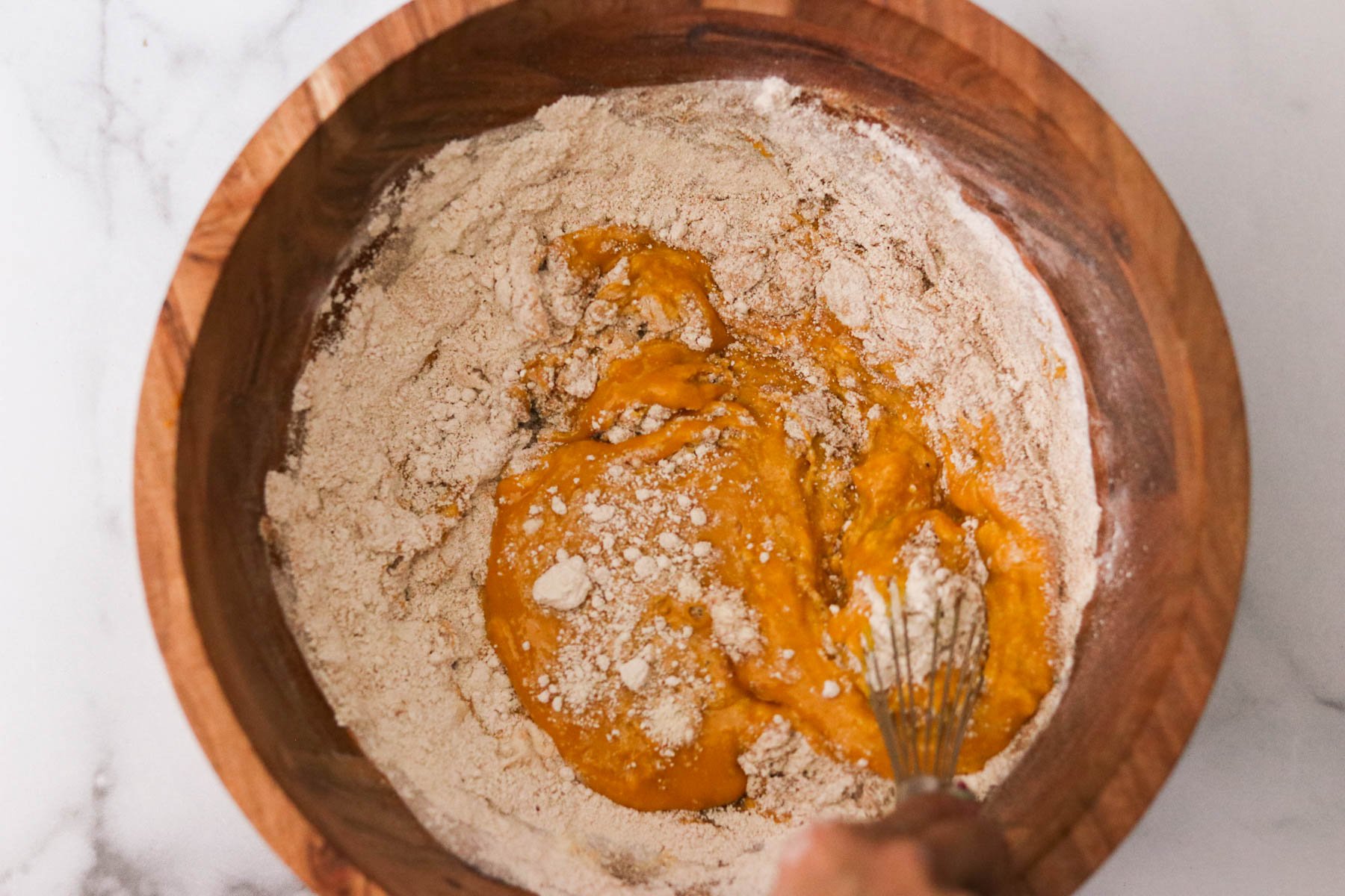 Dry ingredients including oat flour, protein powder, and pumpkin spice added to wet mixture in a wooden mixing bowl.
