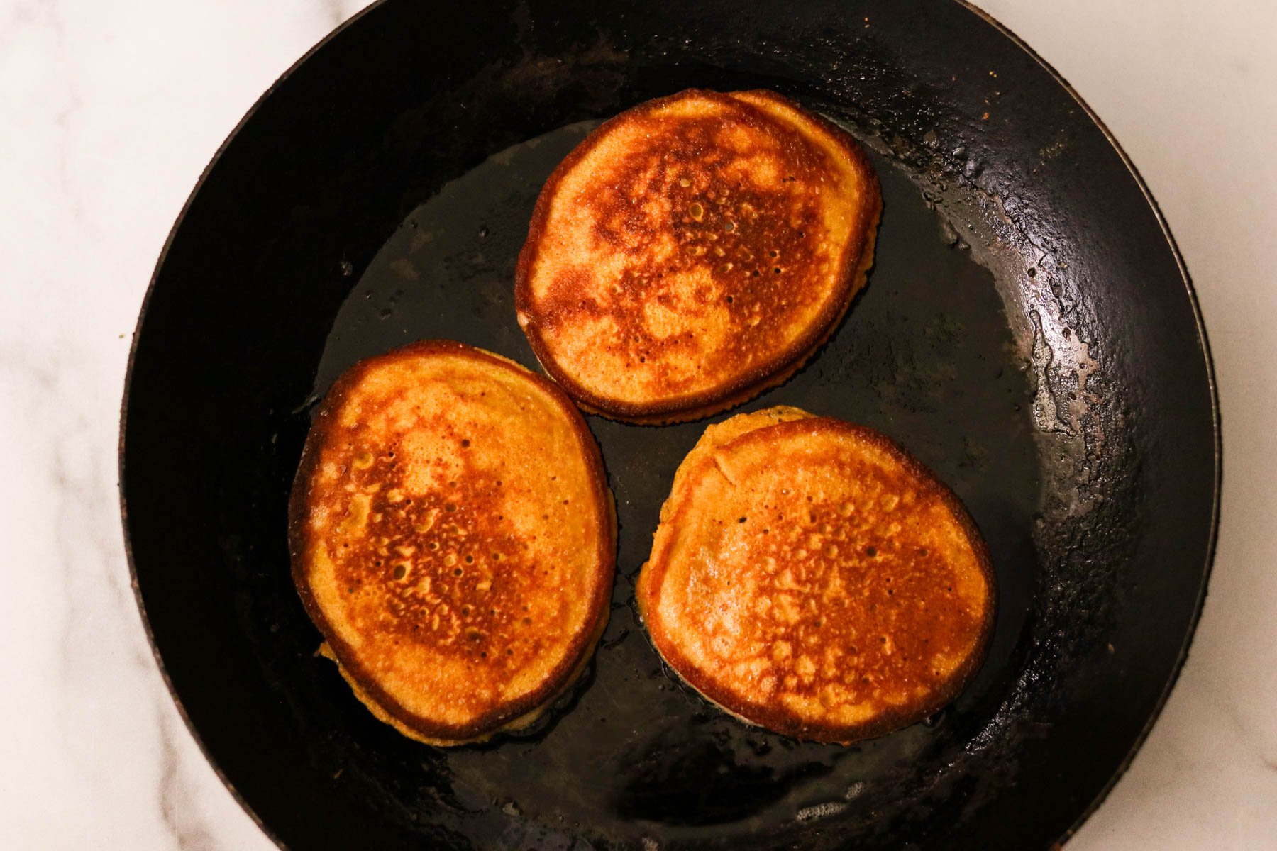 Pumpkin protein pancakes cooking in a black skillet with avocado oil, cooking on medium heat.