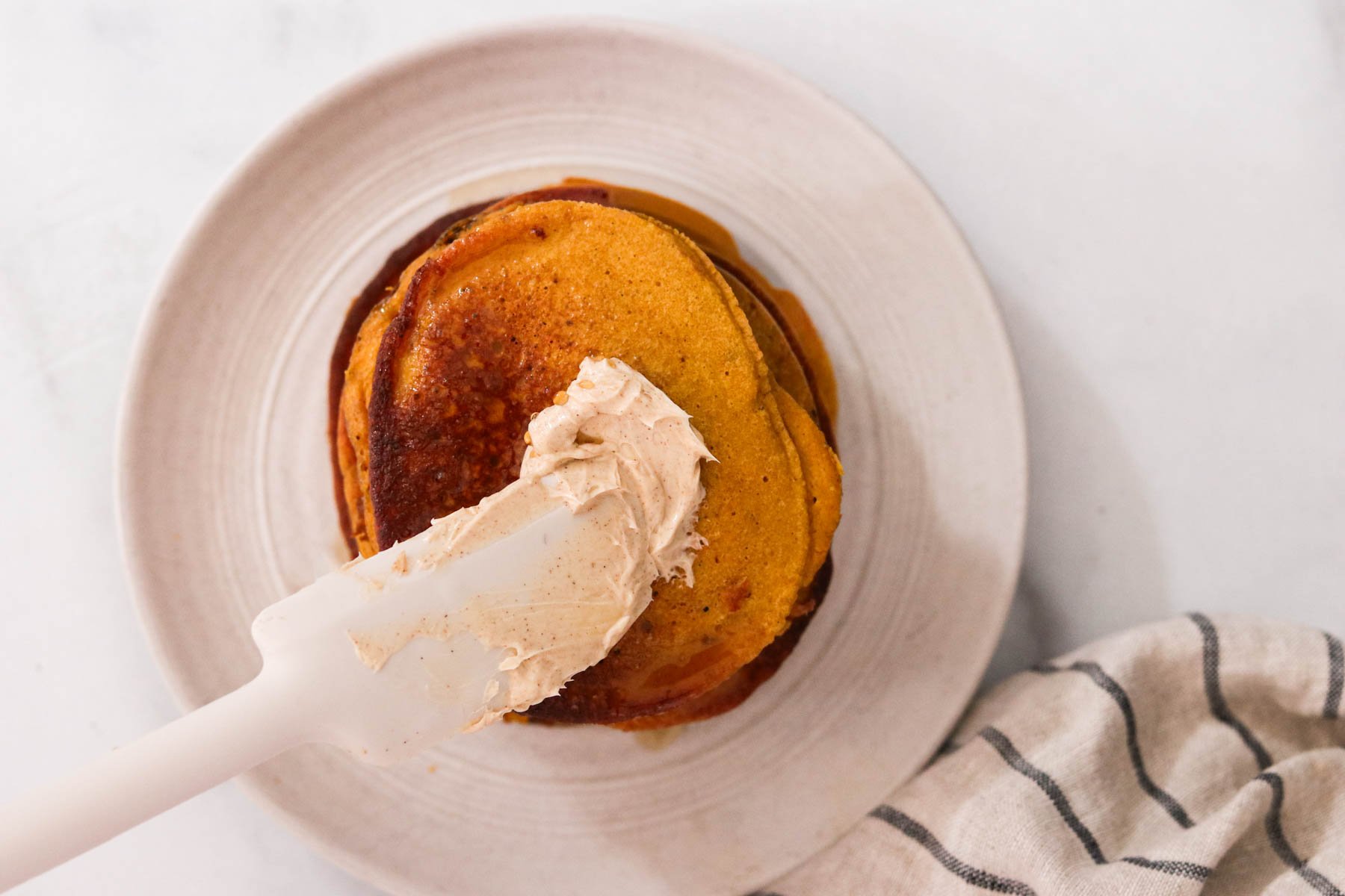 Stack of golden brown pumpkin protein pancakes on a plate with cinnamon maple butter being spread on top, ready for serving with toppings.