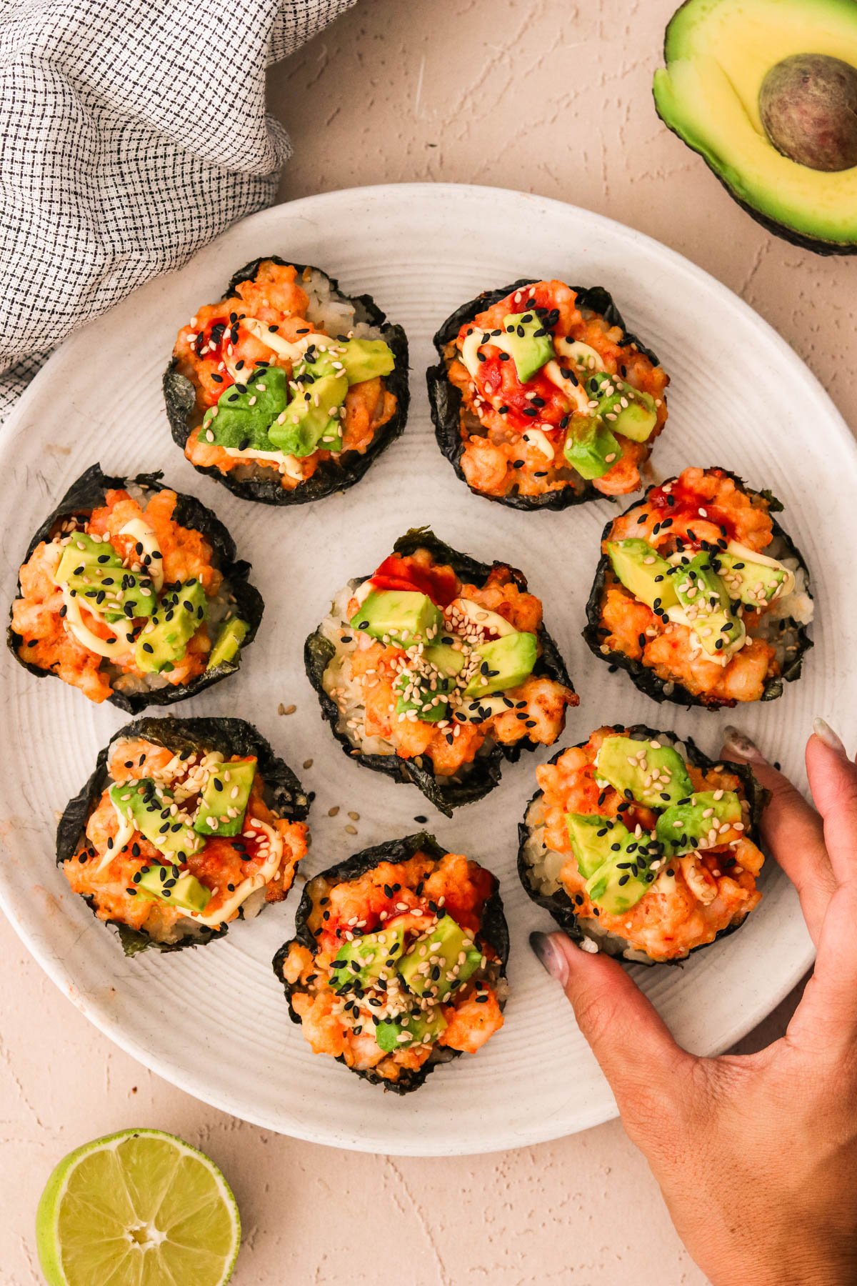 Hand reaching for a shrimp sushi cup topped with avocado and sesame seeds on a white plate.