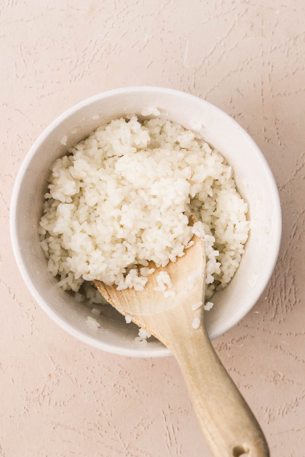 Cooked rice in a small white bowl with a wooden spoon on a light colored surface.