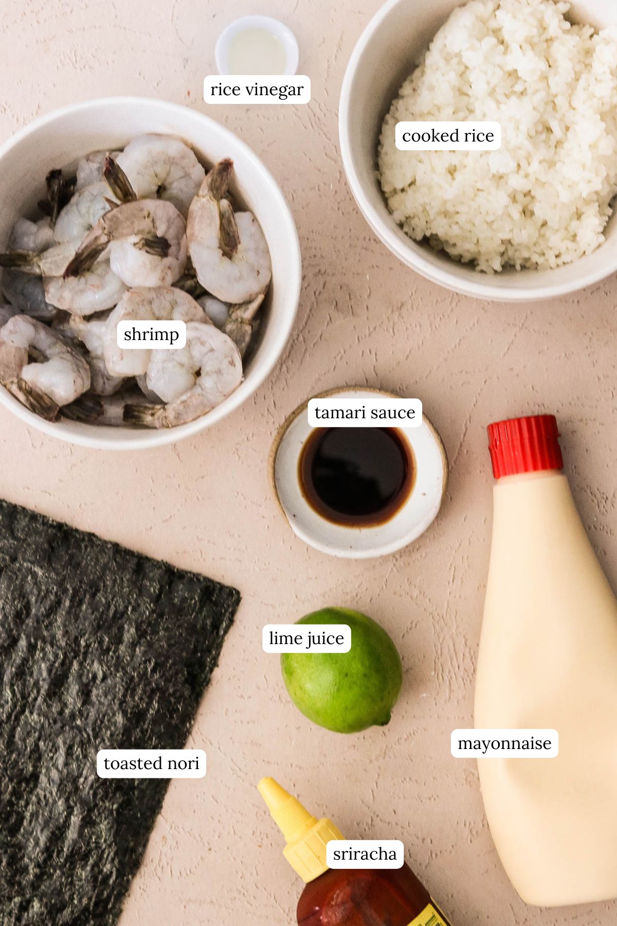 Overhead shot of sushi cup ingredients including cooked rice, nori sheets, shrimp, and seasonings on a light colored kitchen counter.