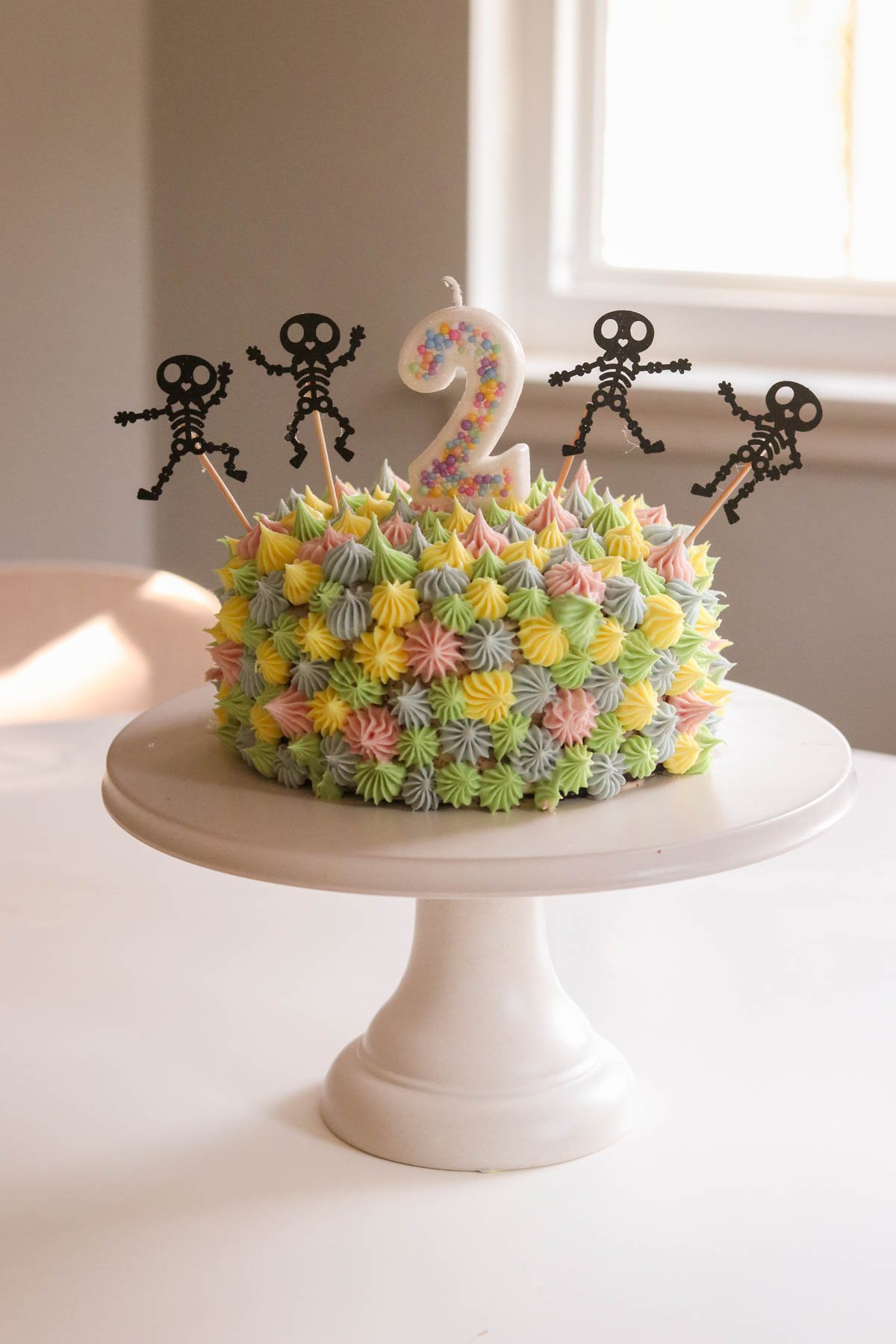 6 inch chocolate birthday cake on a cake stand, decorated with rainbow frosting with skeleton decorations sticking out of it