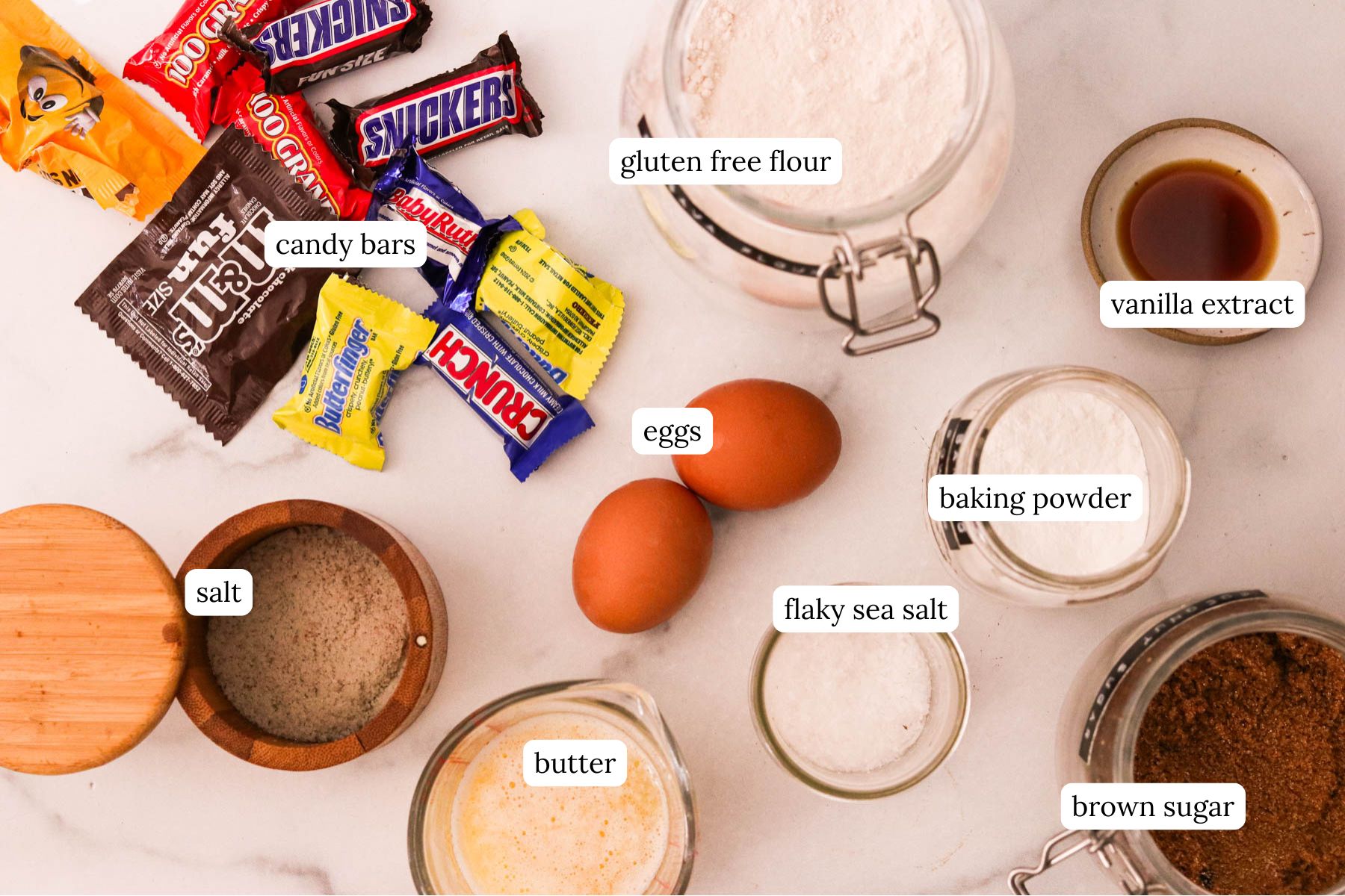 Ingredients for leftover Halloween candy cookie bars including candy bars, gluten free flour, vanilla extract, baking powder, flaky sea salt, eggs, salt, butter, and brown sugar.