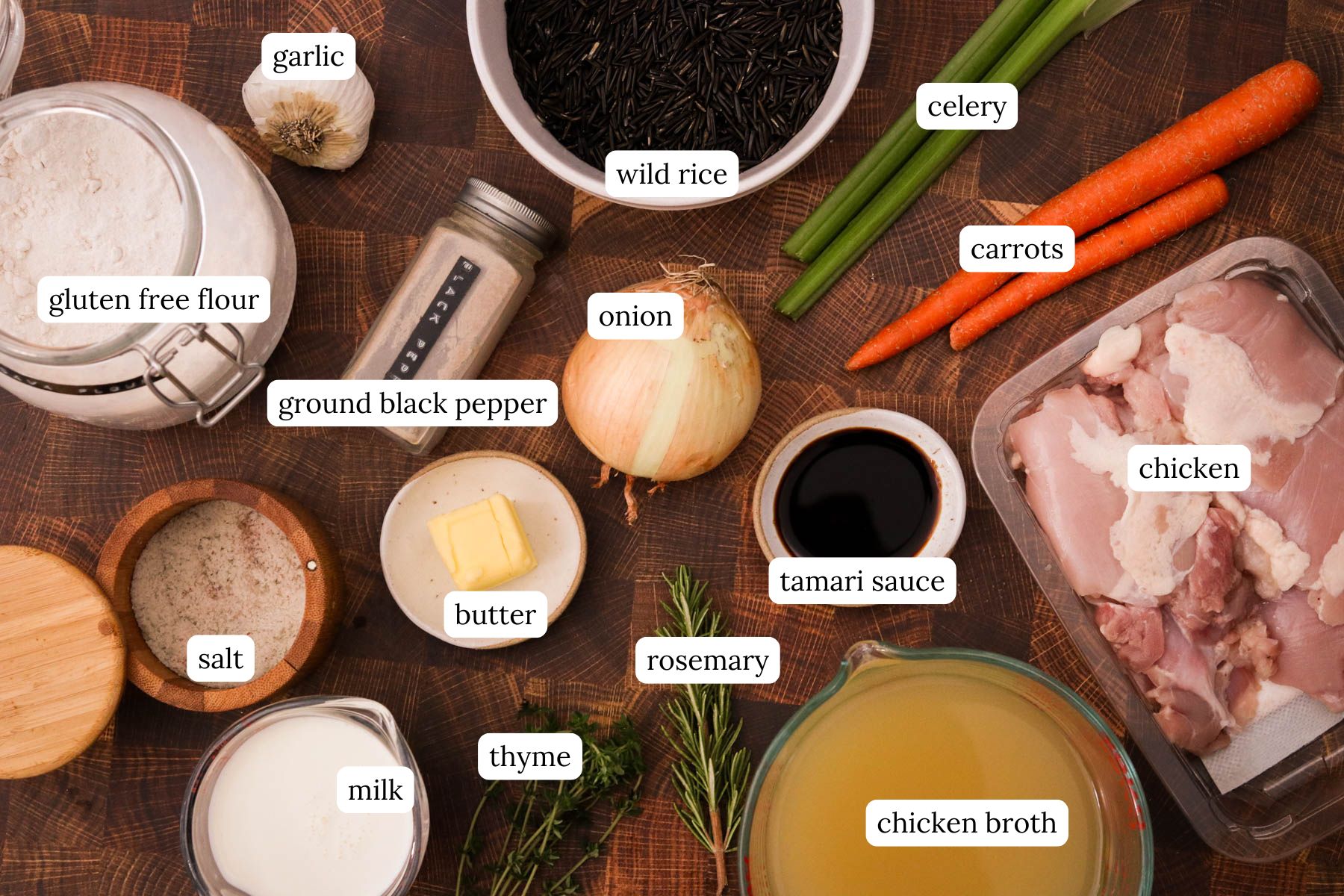 Ingredients for chicken and wild rice soup laid out on a wooden cutting board including wild rice, celery, carrots, chicken, chicken broth, herbs, spices, onion, gluten free flour, garlic, milk, and tamari sauce.