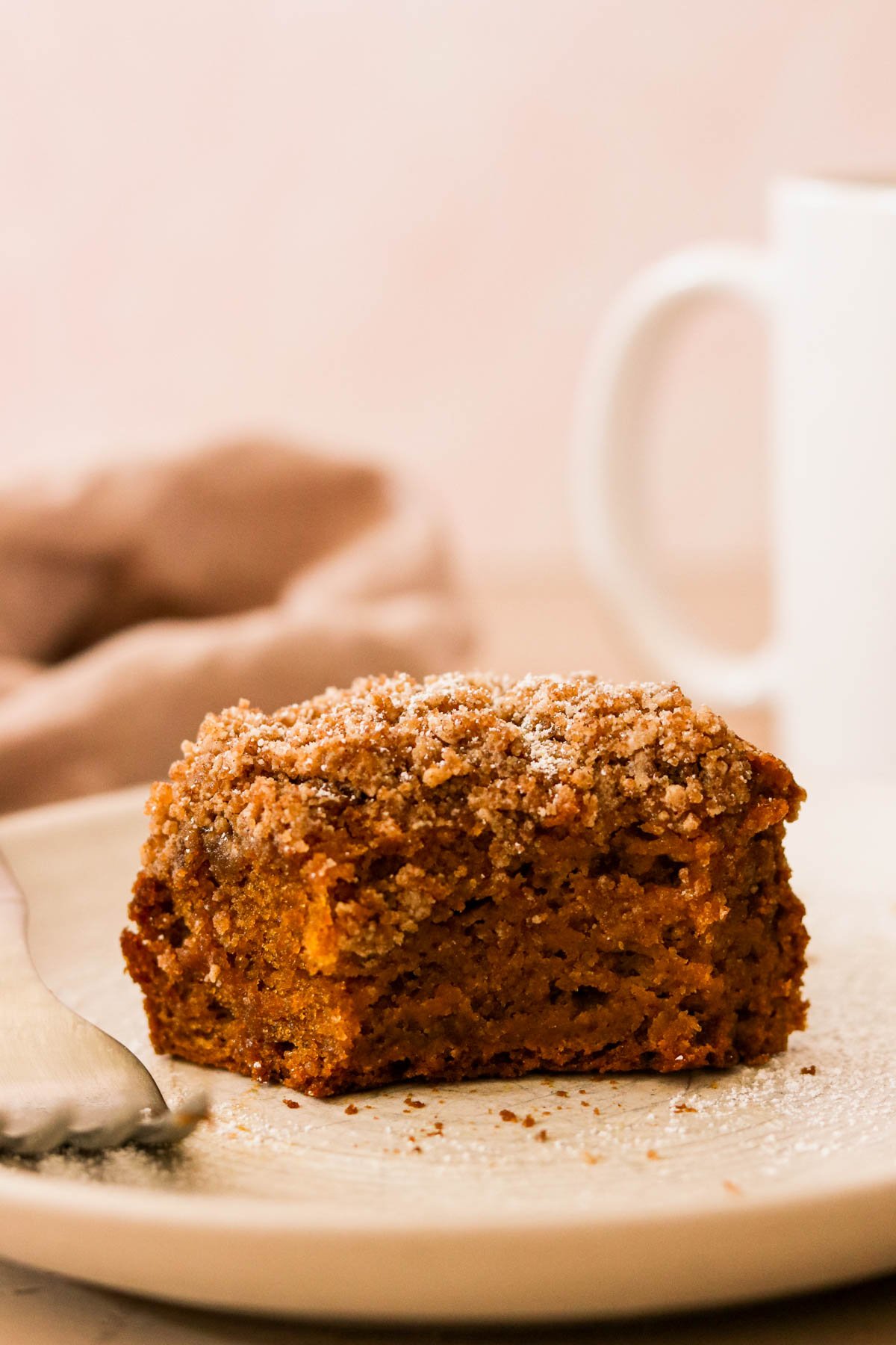 A slice of pumpkin coffee cake with cinnamon crumb topping on a plate, a bite taken out, in front of a light pink backdrop.