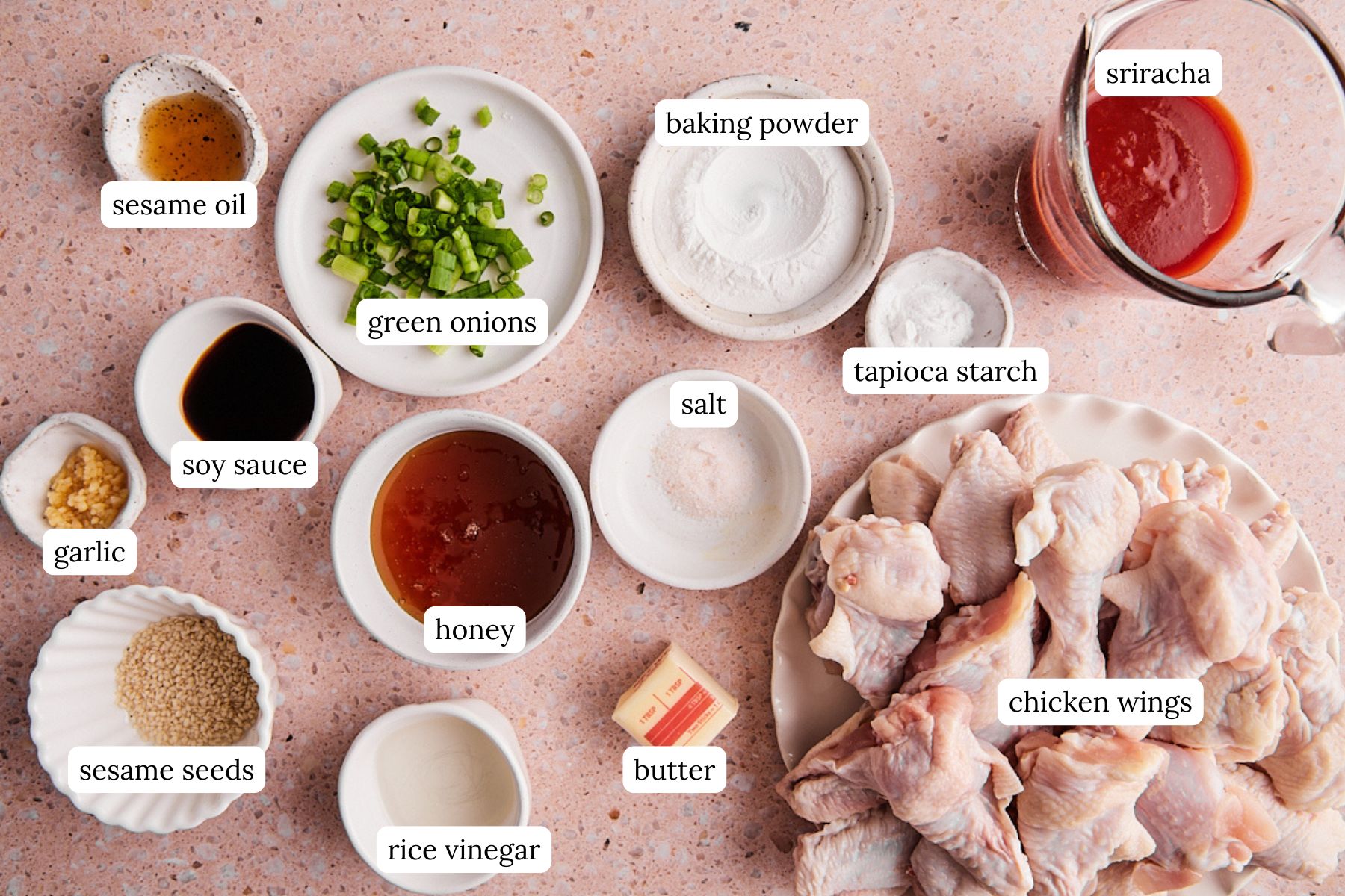 Ingredients for baked honey sriracha wings labeled and laid out on a light pink speckled countertop.