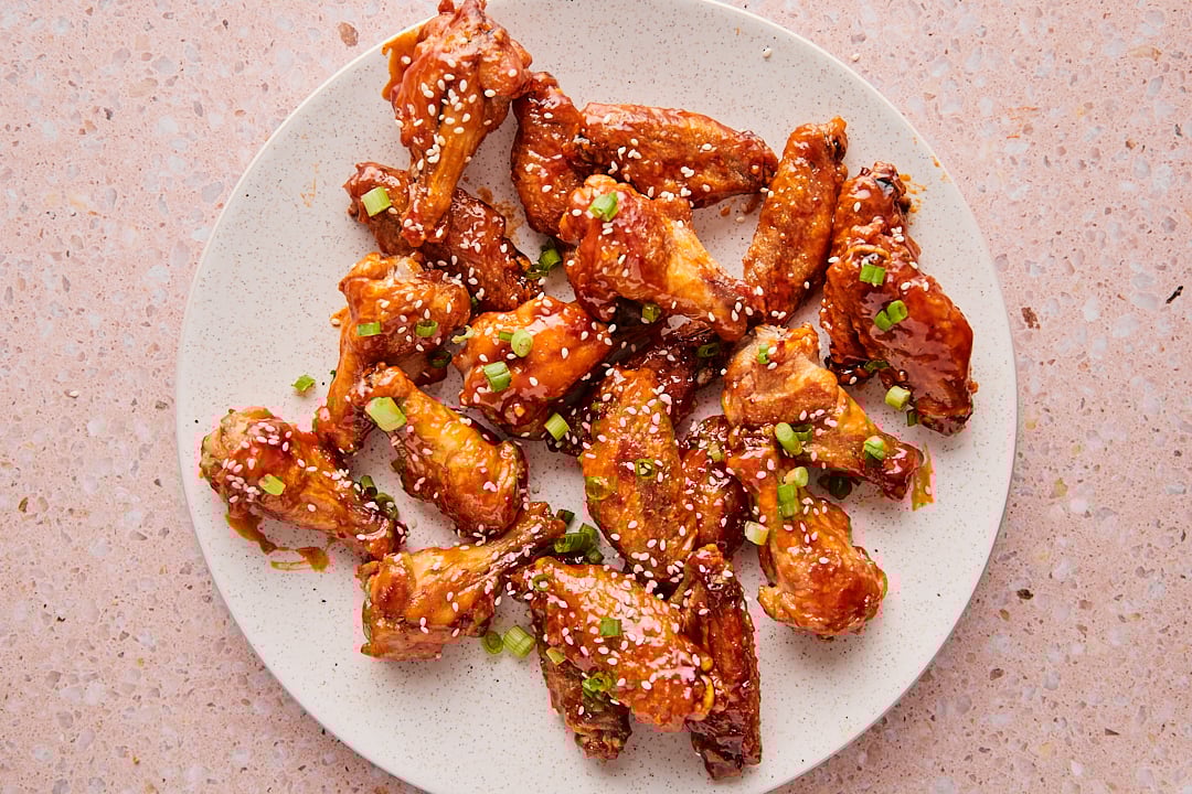 Baked honey sriracha chicken wings on a plate, topped with green onions and sesame seeds, set on a light pink countertop.