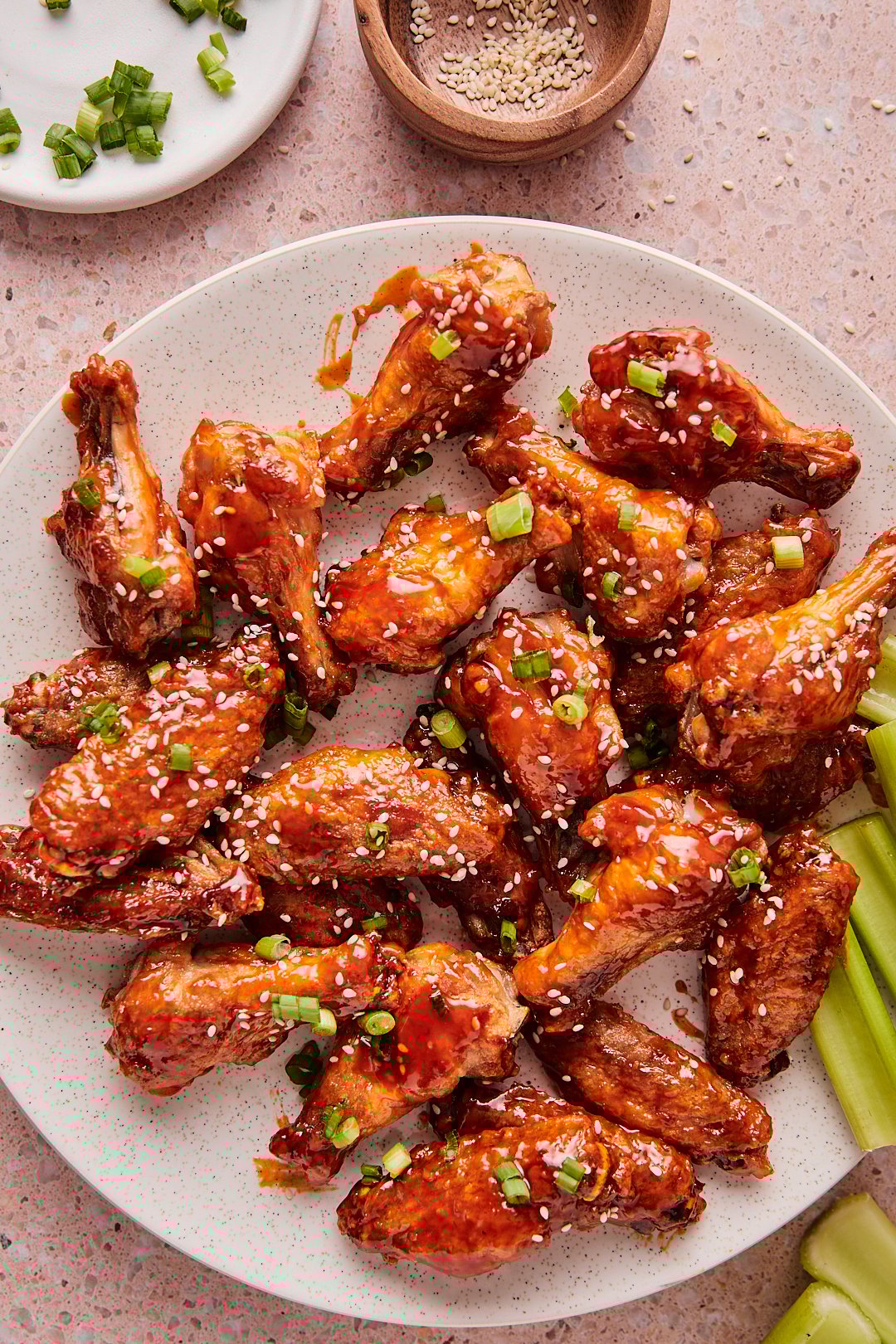 Baked honey sriracha chicken wings on a plate, topped with green onions and sesame seeds with celery on the side, set on a light pink countertop.