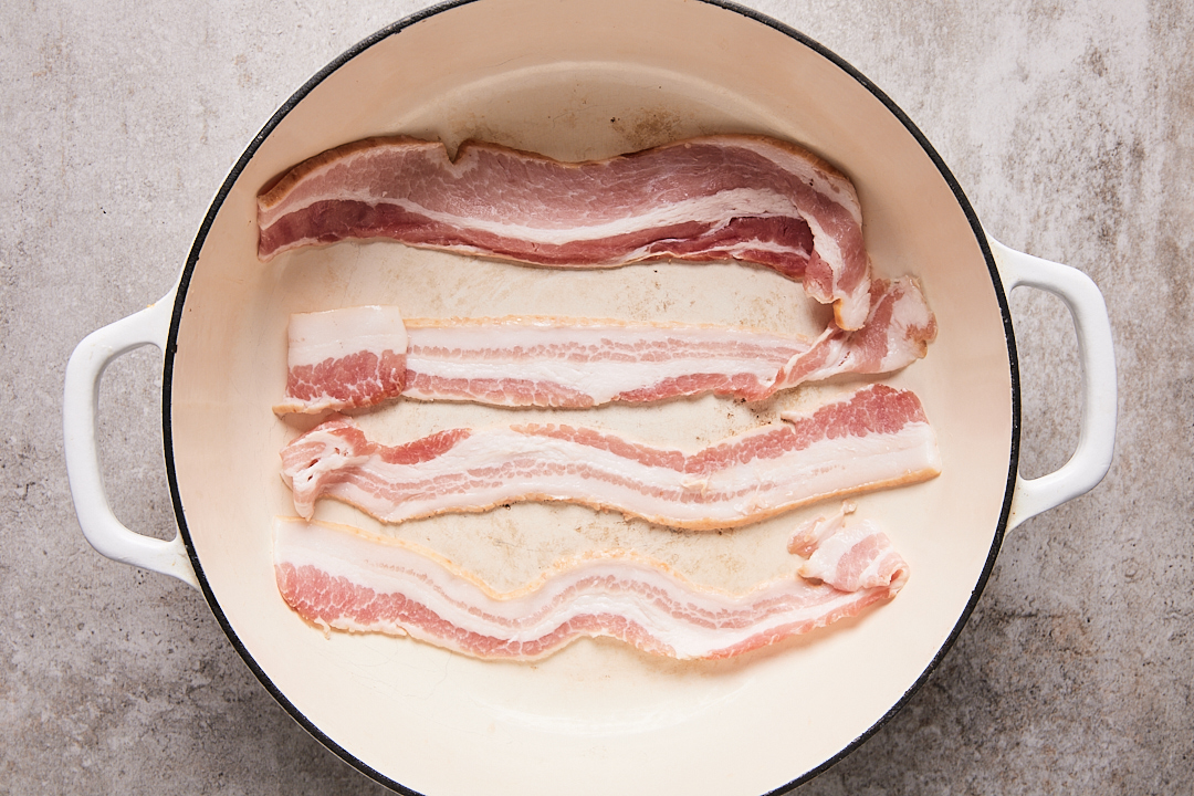 White Dutch oven with raw bacon in it, ready to cook.