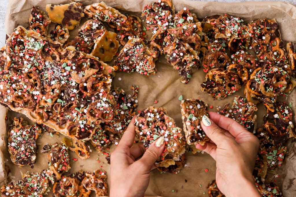 hands breaking christmas crack with pretzels on a parchment lined baking sheet. 