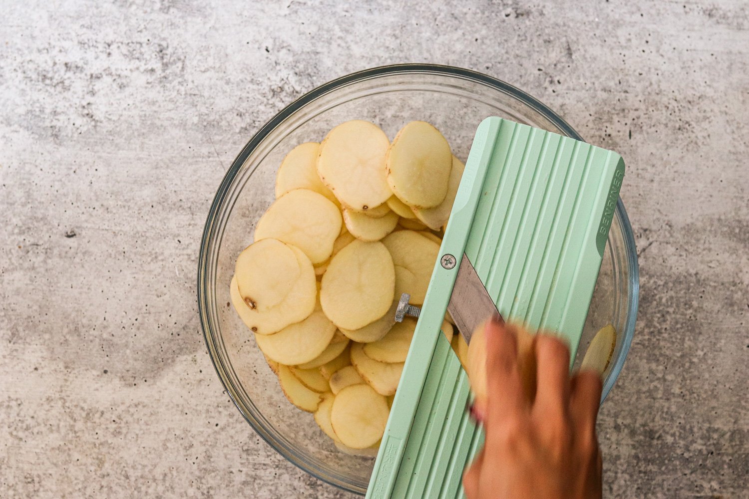 Mandoline slicing 1/8-inch potato rounds into a glass bowl. 