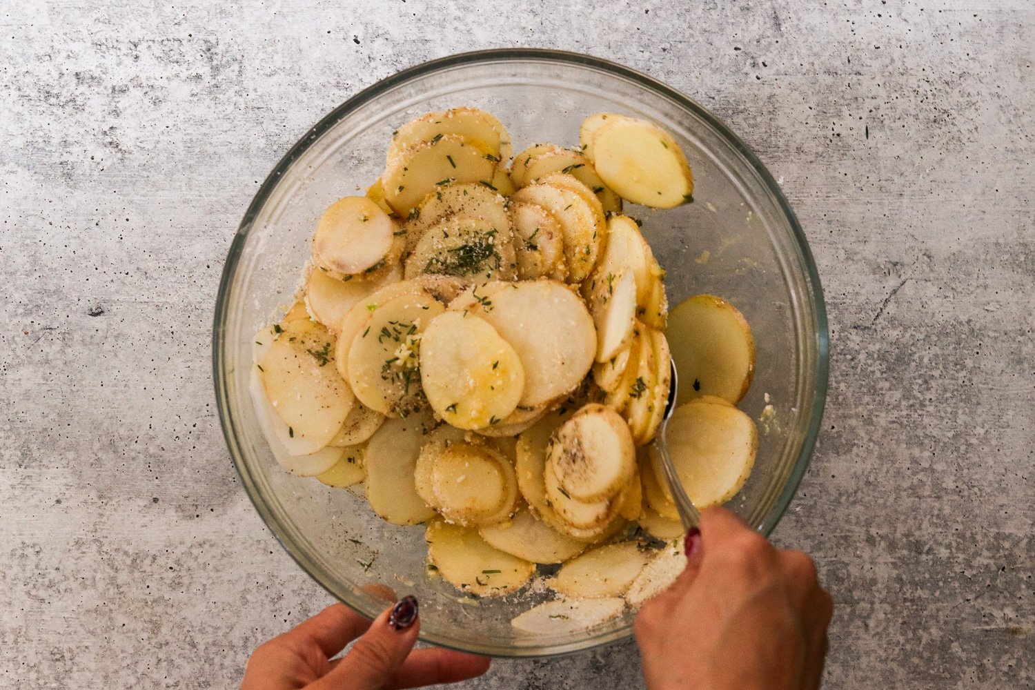Sliced potato rounds in a glass bowl tossed with melted butter, garlic, thyme, rosemary, salt, pepper, and part of the parmesan.
