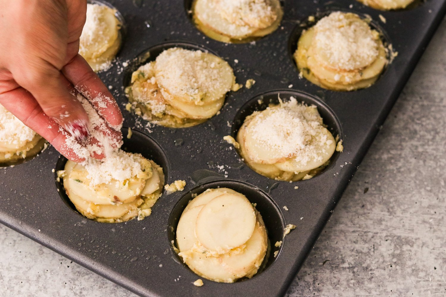 A hand sprinkling Parmesan cheese on top of the potato stacks in a muffin tin.