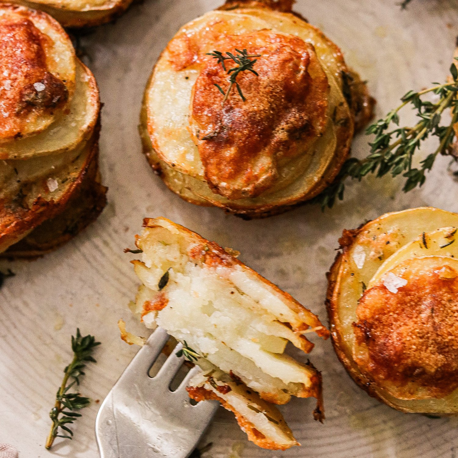 Plated crispy stacked potatoes garnished with flaky sea salt and fresh thyme, with one potato cut open and resting on a fork, ready to be eaten.