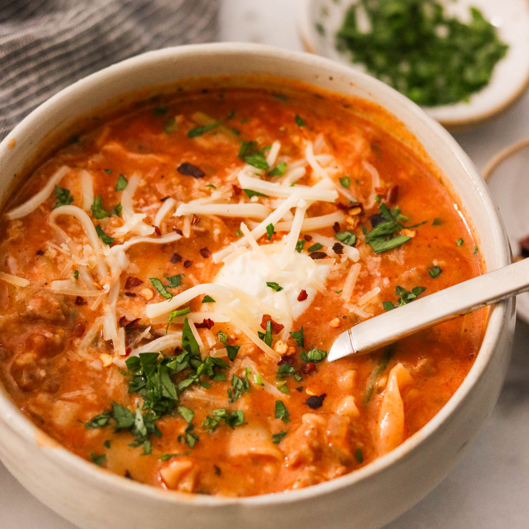 Close-up of Instant Pot lasagna soup in a bowl, topped with cheeses and fresh herbs, with a spoon ready to eat.
