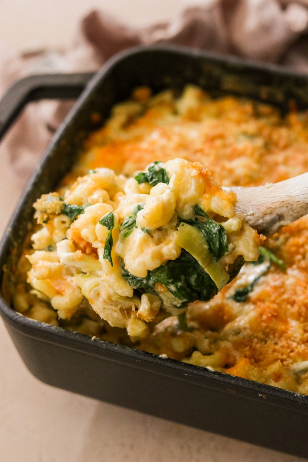 A wooden spoon scooping out a portion of spinach and artichoke mac and cheese from a black 9x13 STAUB pan.