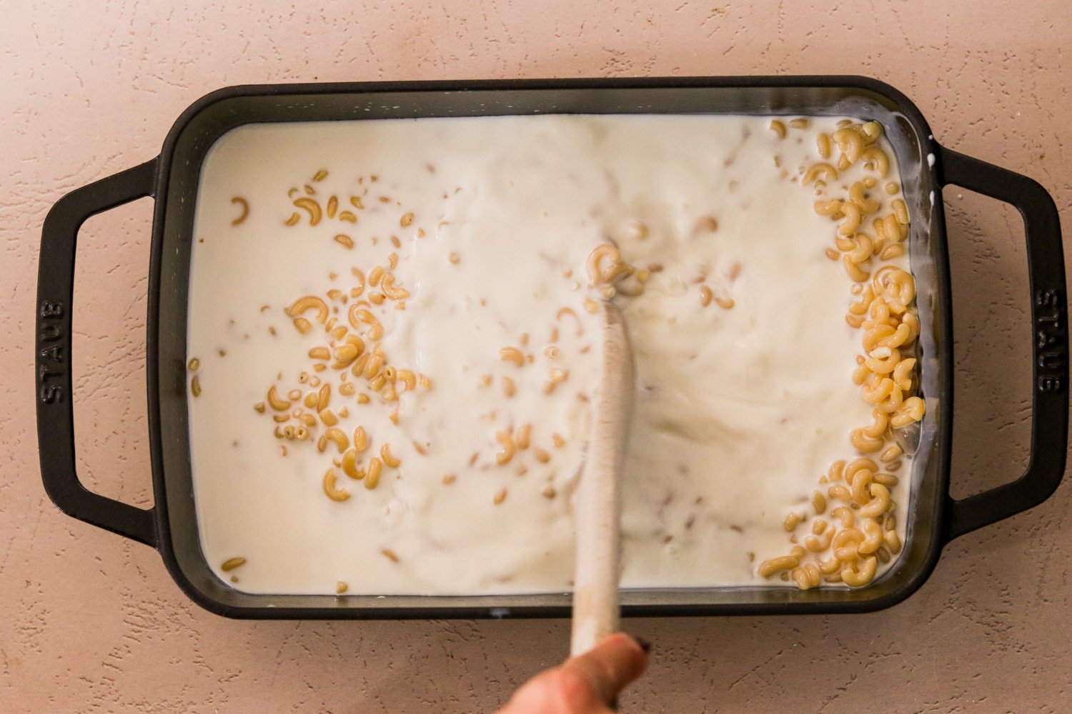 Pasta, milk, and sea salt in a 9x13 pan being stirred with a wooden spoon.