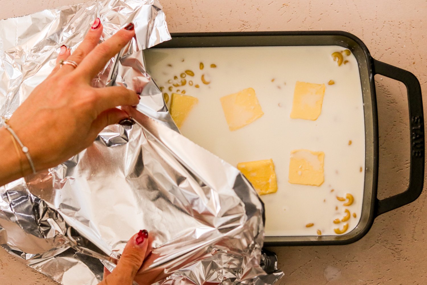 Hands covering a 9x13 baking dish filled with pasta, milk, sea salt, and sliced butter with aluminum foil.