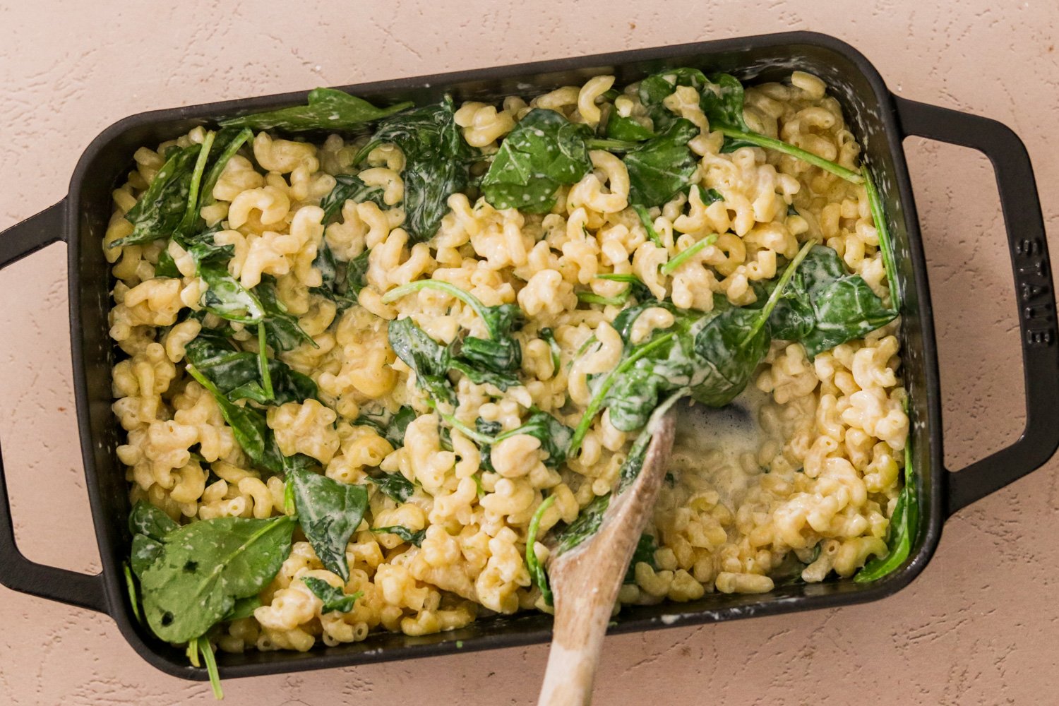 A wooden spoon breaking up baked spinach and artichoke mac and cheese in a 9x13 pan.