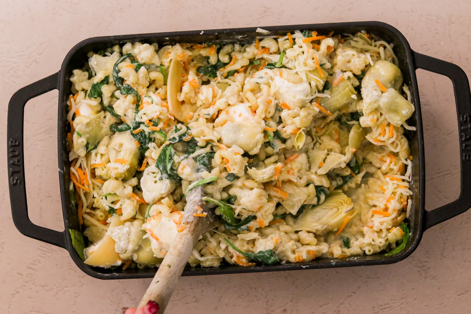 Baby spinach and artichoke hearts being stirred into the 9x13 pan with baked mac and cheese.