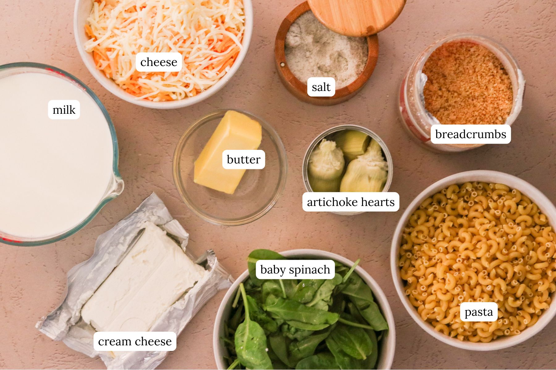Ingredients for spinach and artichoke mac and cheese labeled and laid out on a light pink countertop. 